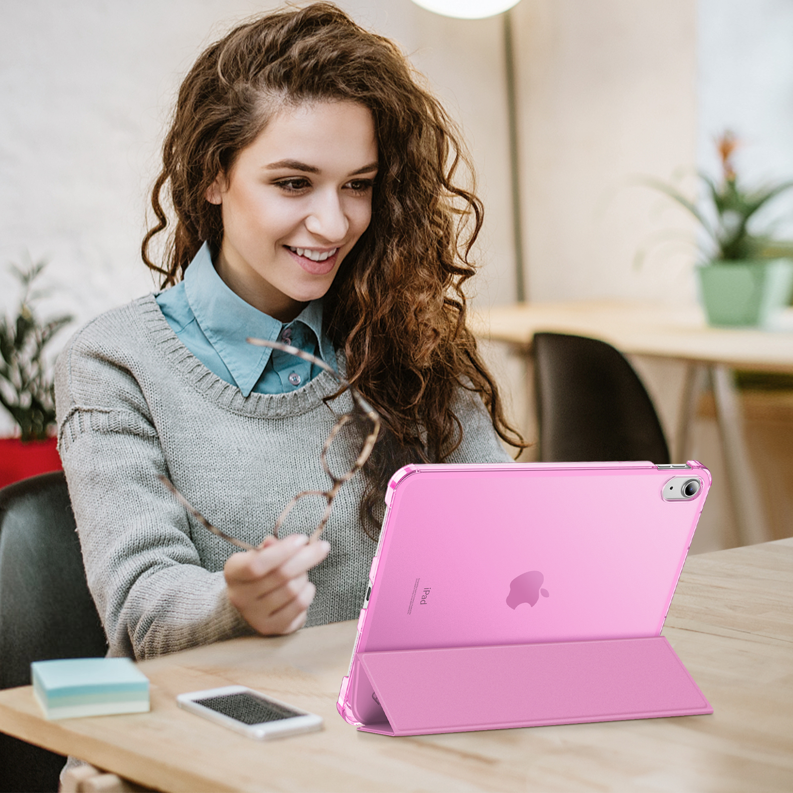 Eine lächelnde Frau hält eine Brille und schaut auf ein pinkfarbenes iPad. Auf dem Tisch liegen ein Telefon und Haftnotizen.