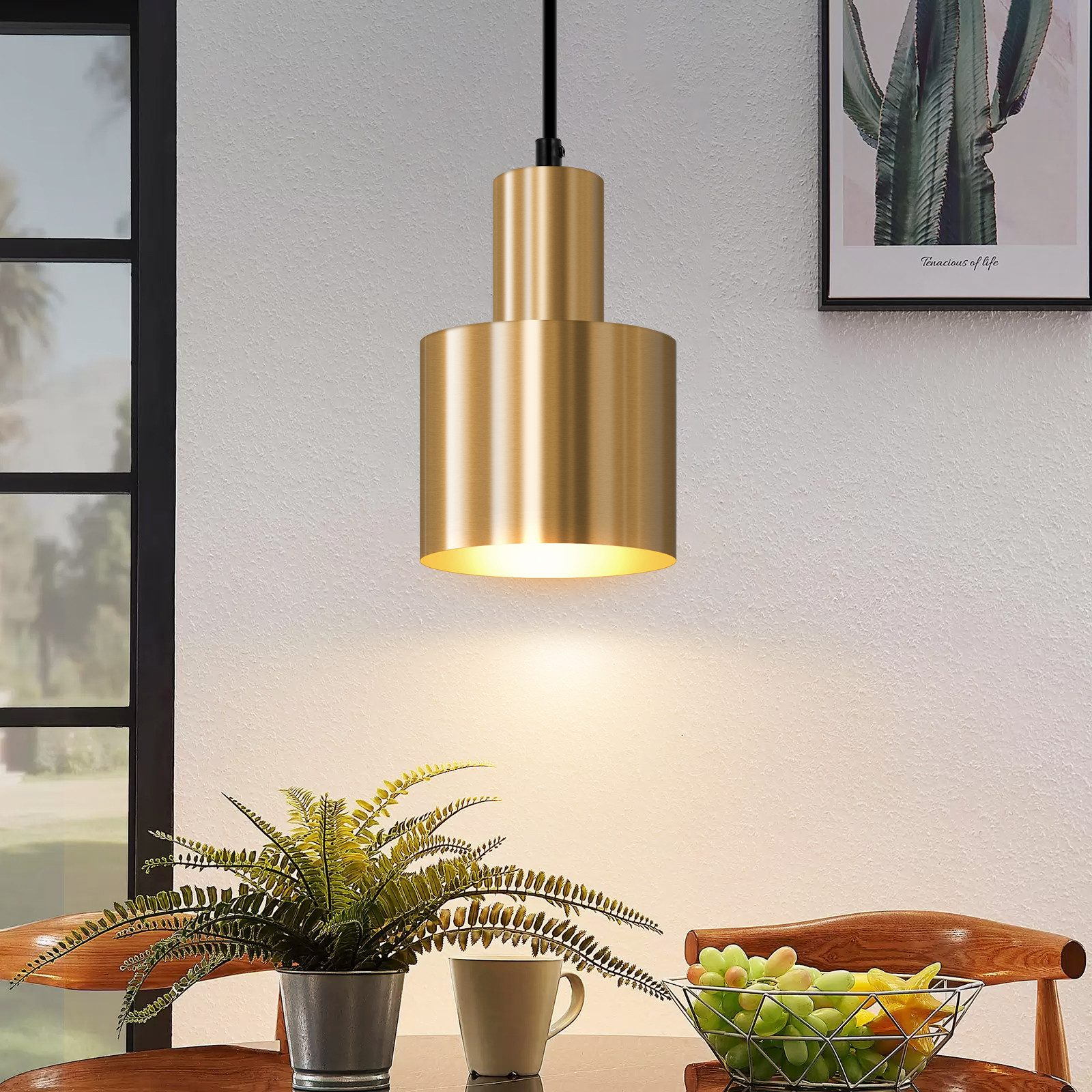 Eine goldene Pendelleuchte hängt über einem Tisch mit einer Pflanze, einer Tasse und einem Obstkorb. Ein Kaktus-Kunstwerk an der Wand.