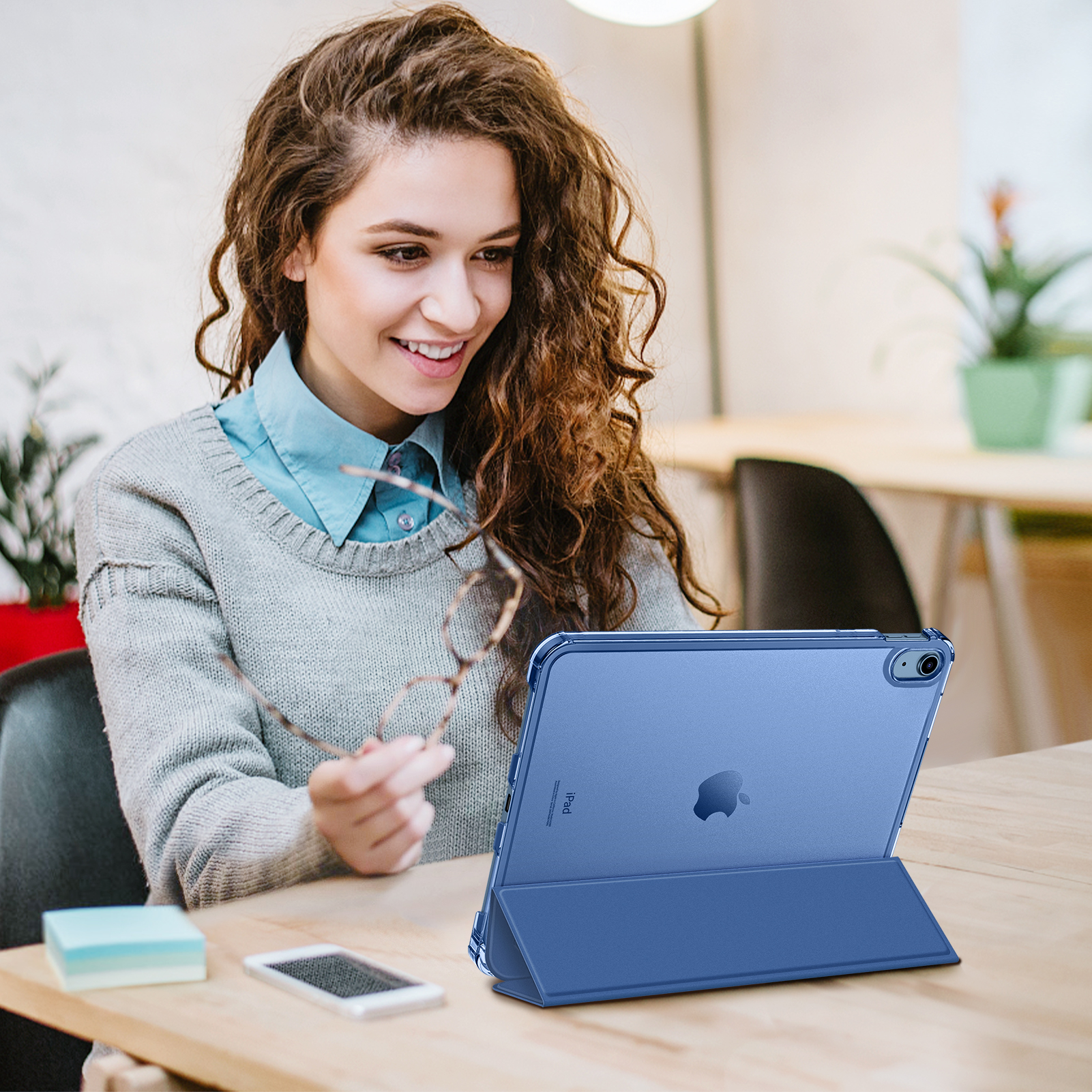 Eine Frau lächelt und hält eine Brille. Sie schaut auf ein blaues iPad auf einem Tisch. Sie trägt einen grauen Pullover und ein blaues Hemd.