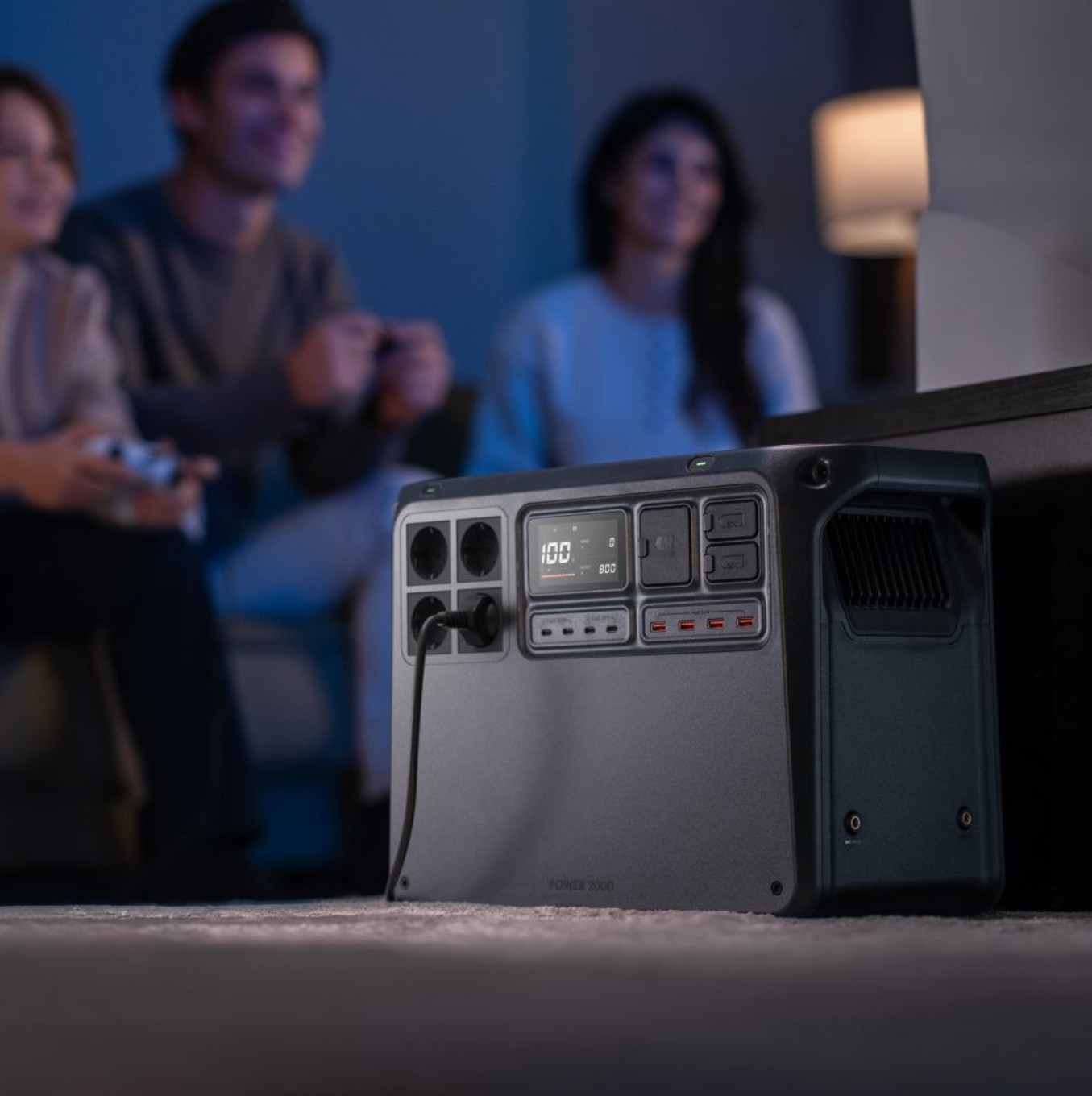 Eine Powerstation ist angeschlossen. Im Hintergrund sind drei Personen verschwommen, die ein Spiel spielen, eine Person hält einen Controller.