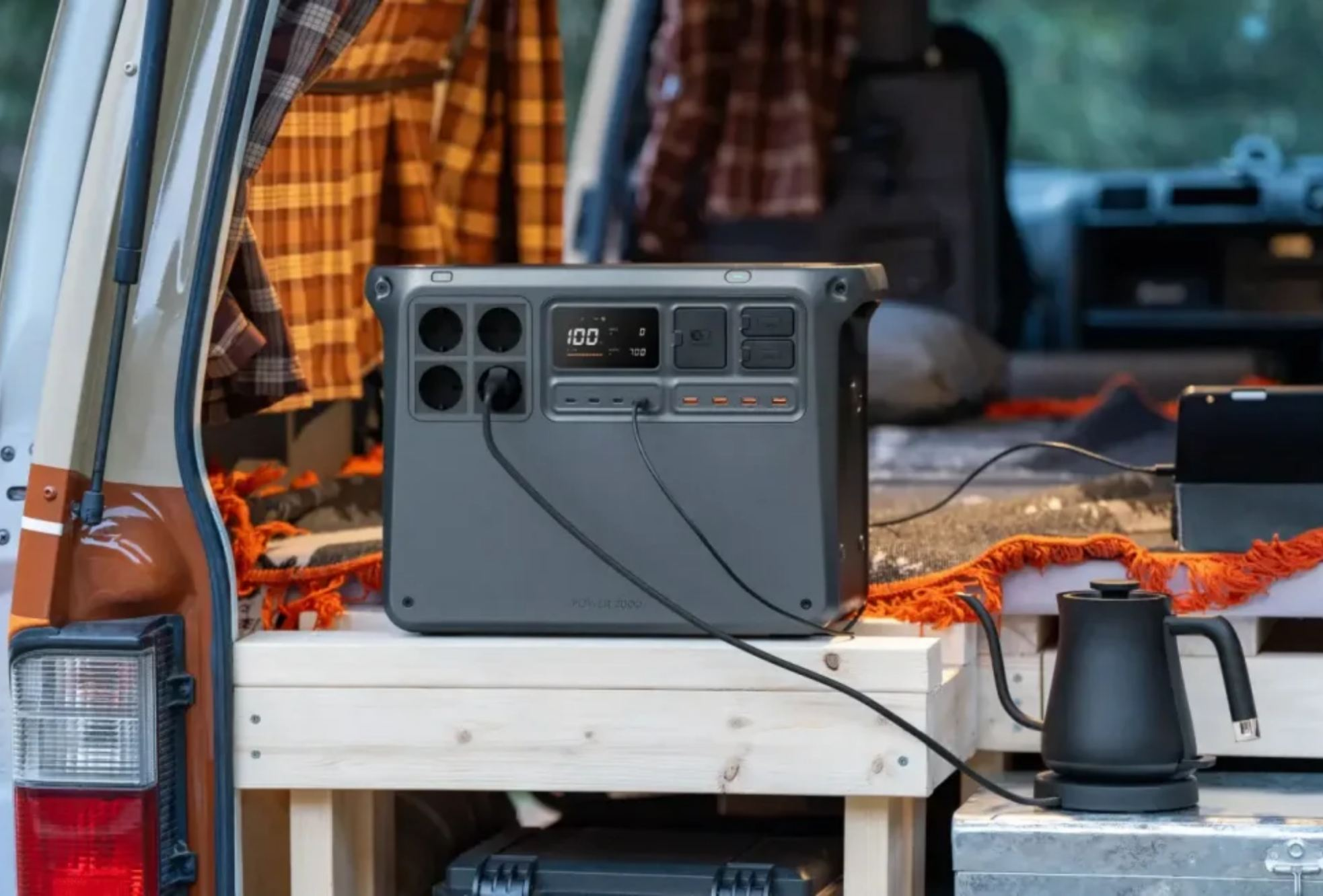 Graue Powerstation in einem Van, mit Wasserkocher und Tablet. Orangefarbene Decke auf der Holzplattform. Karierte Vorhänge sind sichtbar.