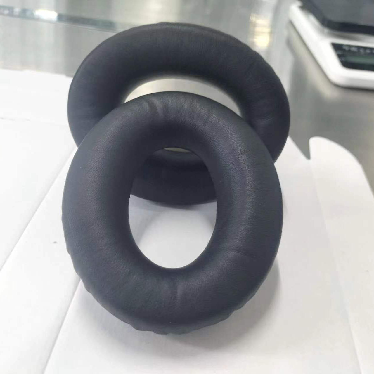 Due cuscinetti auricolari neri e circolari per cuffie, uno sopra l'altro. Sono su una superficie bianca. Una bilancia è visibile sullo sfondo.