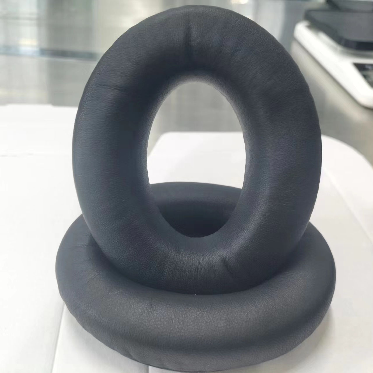 Due cuscinetti auricolari neri a forma di ciambella impilati su una superficie bianca. Quello superiore è leggermente decentrato.