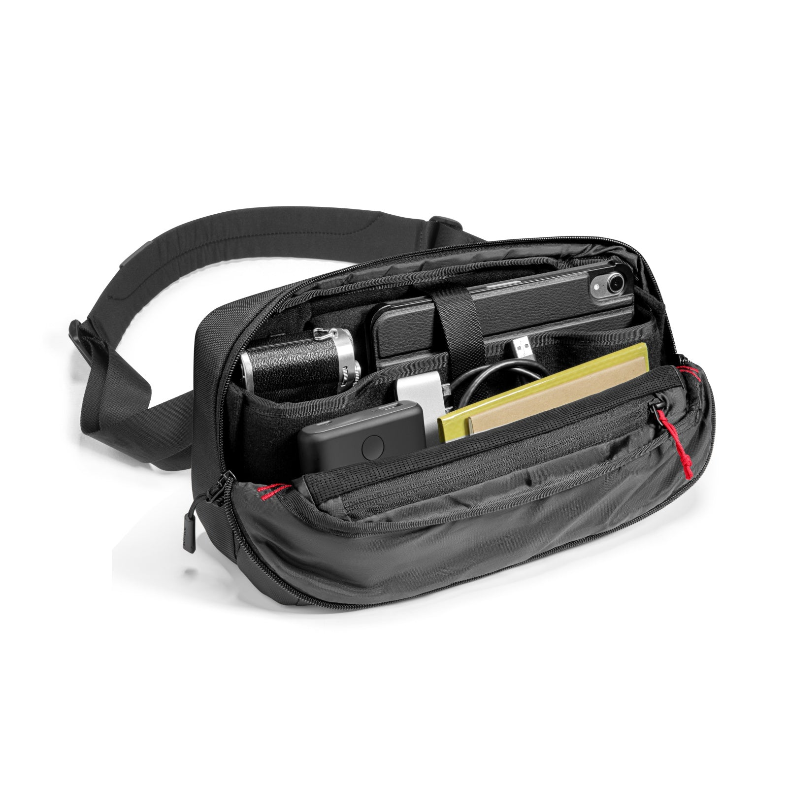 Czarna torba z aparatem, telefonem, ładowarką i notatnikiem w środku. Czerwony zamek błyskawiczny. Czarny pasek na białym tle.