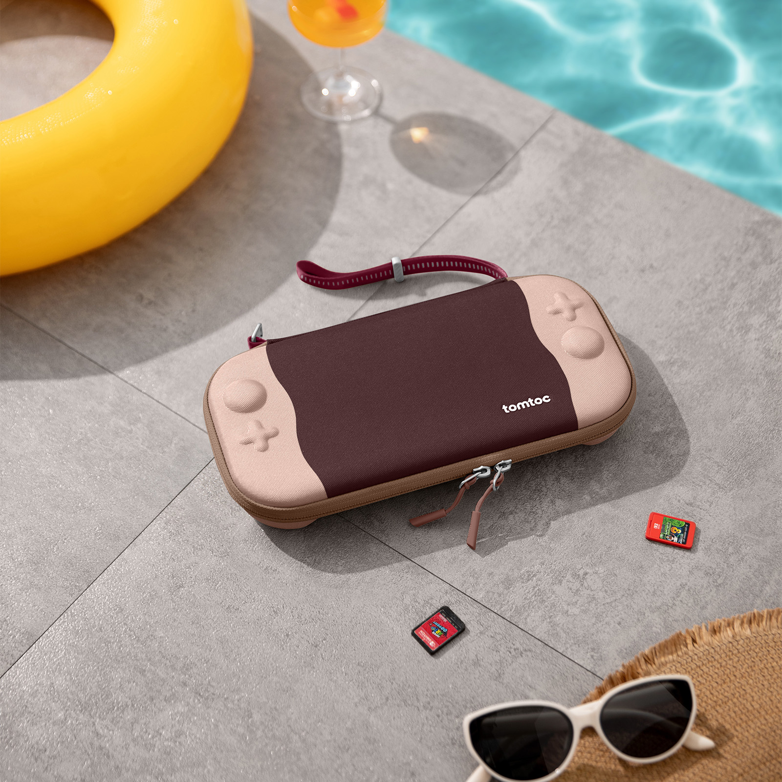 Etui na Switch, dwie kasety do gier, okulary przeciwsłoneczne i kapelusz znajdują się na patio. W pobliżu żółty pływak i napój.