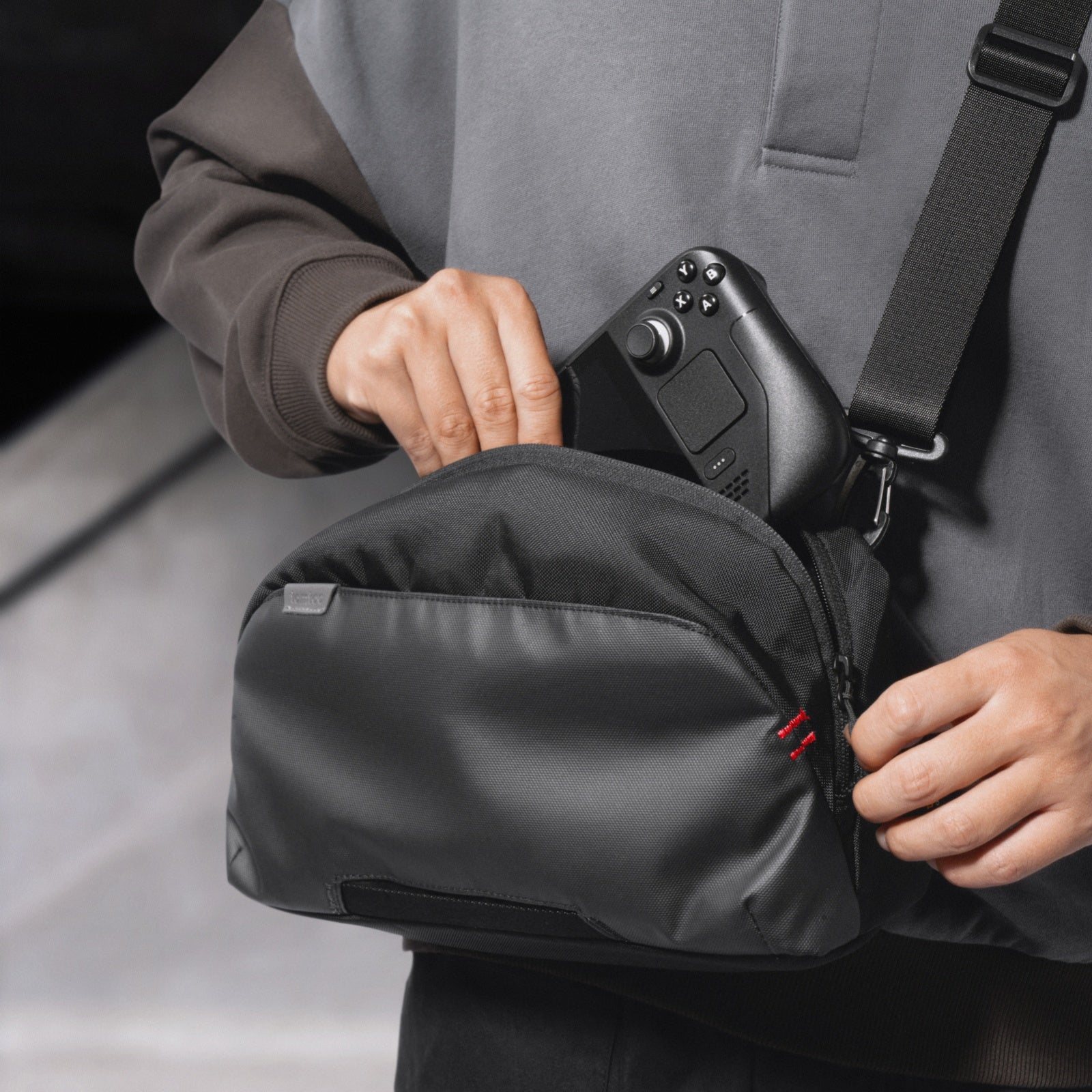 Osoba wkłada czarne, przenośne urządzenie do gier do czarnej torby na ramię. Torba ma czarny pasek i czerwony zamek błyskawiczny.