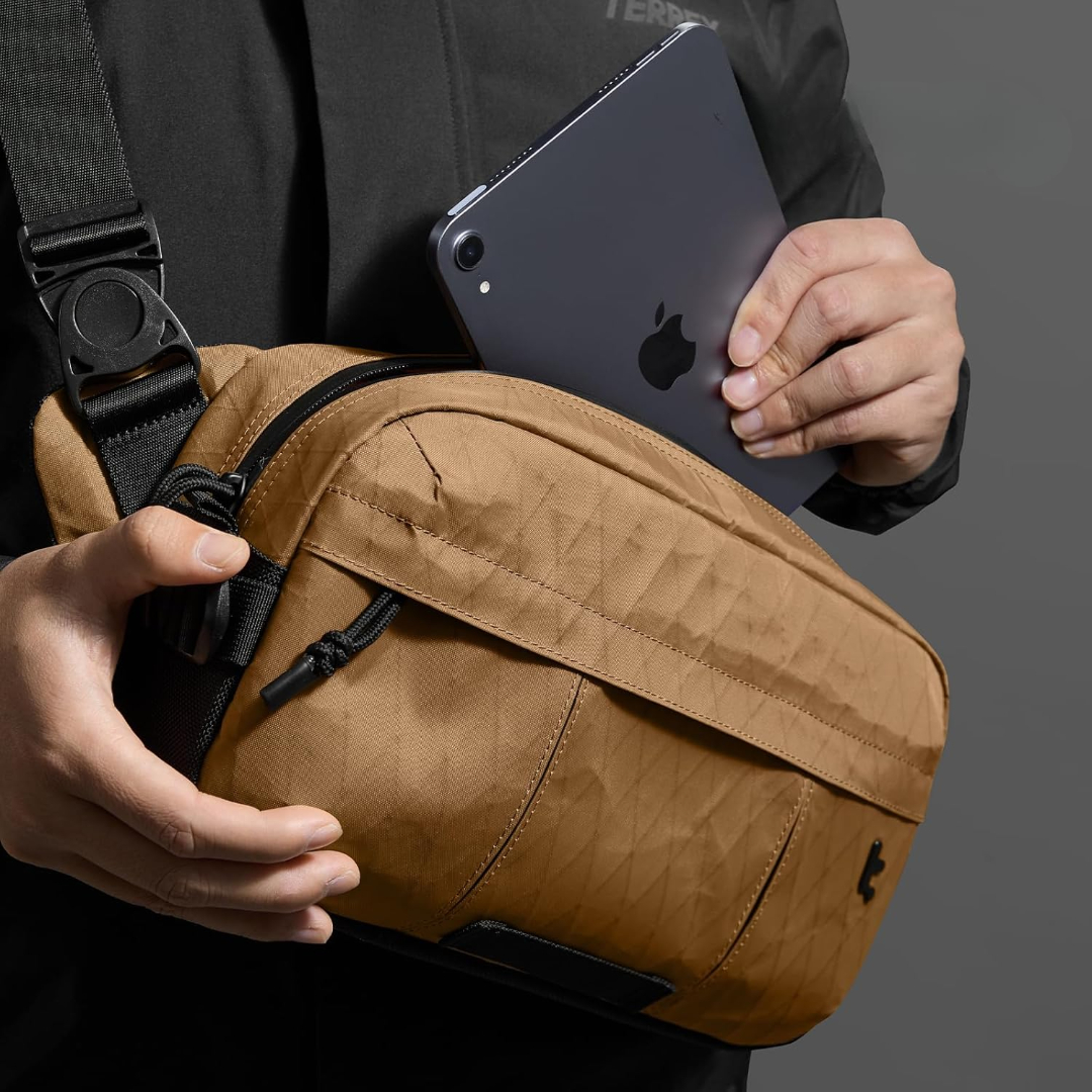 Osoba trzyma tablet nad brązową torbą. Torba ma czarny pasek. Osoba ma na sobie czarną kurtkę. Szare tło.