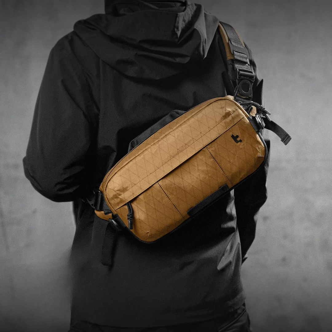 Osoba w czarnej kurtce z brązową torbą na ramię. Torba ma czarny pasek i małe czarne logo.