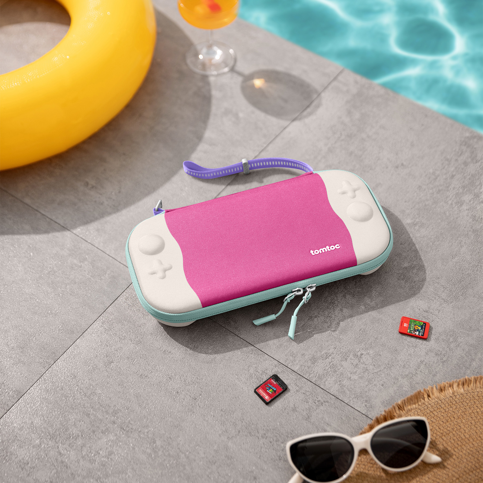 Różowe i białe etui na Switch, w pobliżu basenu z żółtym kołem ratunkowym, okularami przeciwsłonecznymi i kartridżami do gier. Słoneczny dzień.