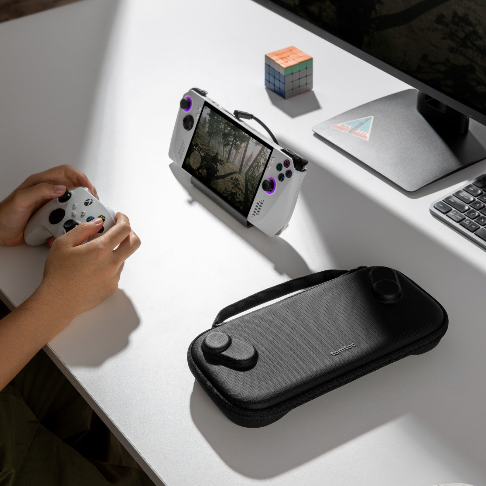 Osoba trzymająca biały gamepad, obok przenośnego urządzenia do gier, etui, kostki Rubika i monitora.