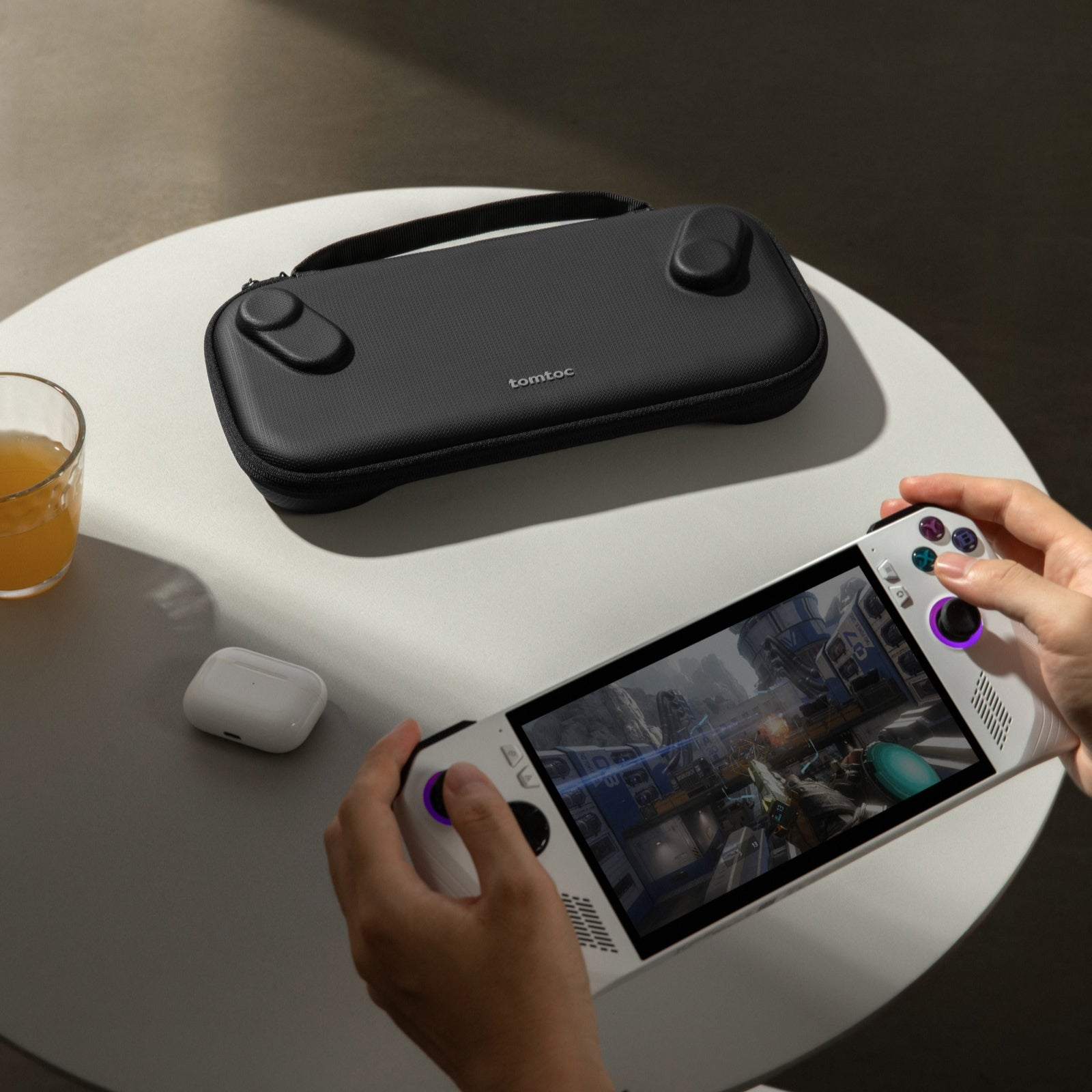 Osoba trzyma urządzenie do gier, etui, szklankę soku i parę białych słuchawek na białym stole.
