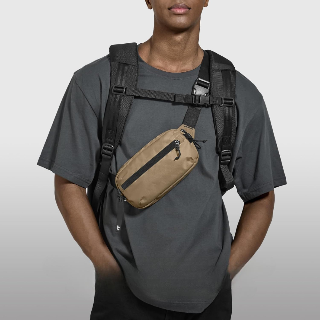 Mężczyzna w szarej koszulce, czarnym plecaku i beżowej nerce z czarnym paskiem. Jest na tle białego tła.