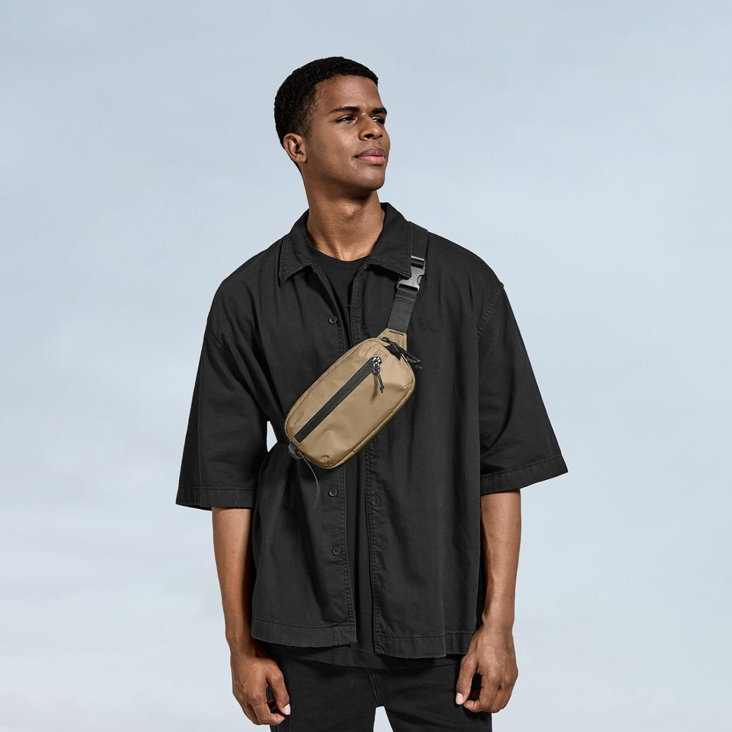 Mężczyzna w czarnej koszuli i spodniach nosi beżową nerkę. Jasnoniebieskie tło. Mężczyzna patrzy w lewo.