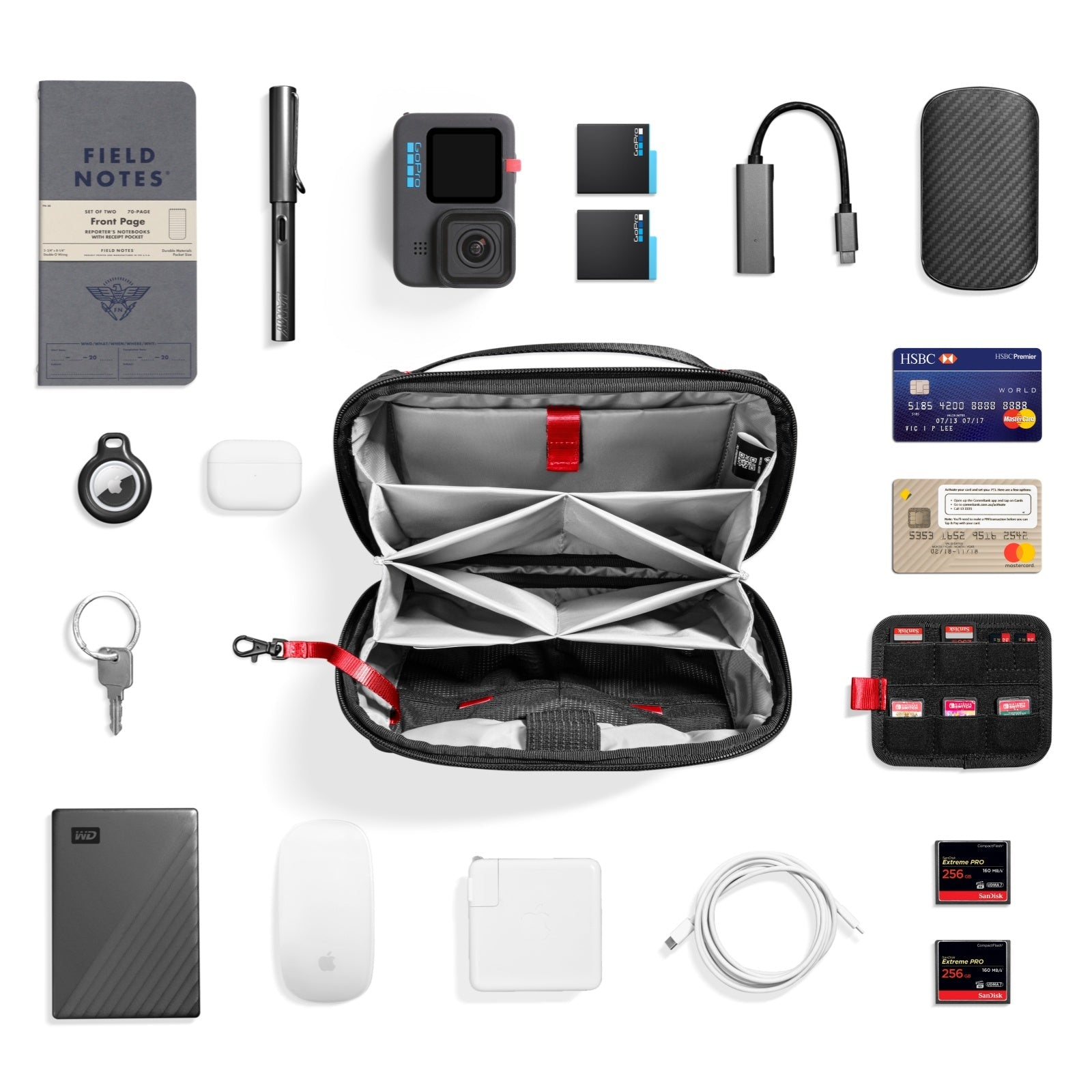 Widok z góry na przedmioty techniczne: notatnik, długopis, aparat, baterie, adapter, etui, karty, AirTag, AirPods, klucze, dysk, mysz, ładowarka, kabel i karty SD.