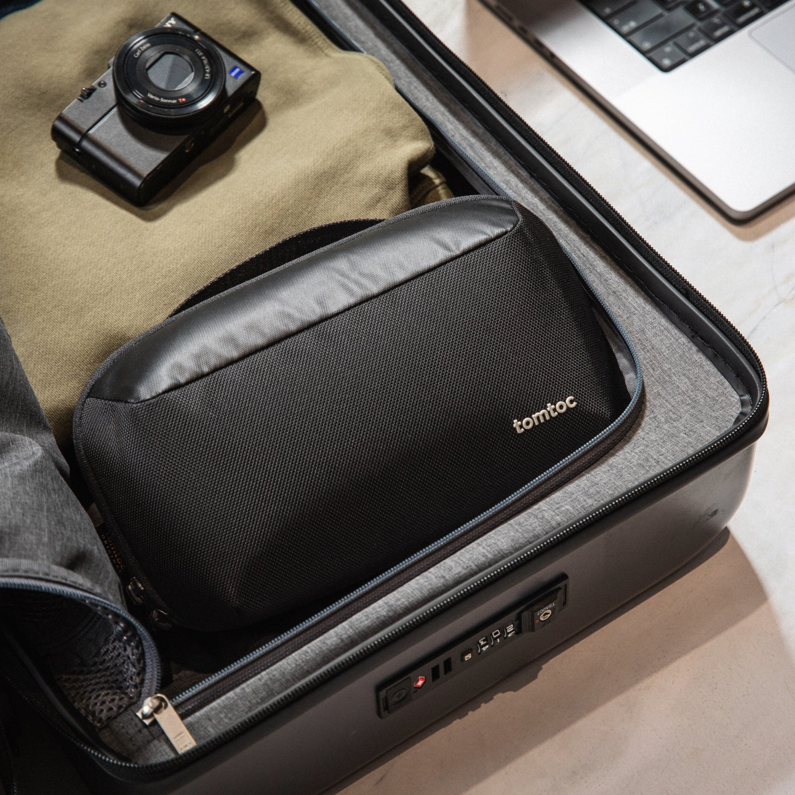 Otwarta walizka z czarnymi akcesoriami: aparat, torba i laptop. Torba z napisem 'tomtoc'.
