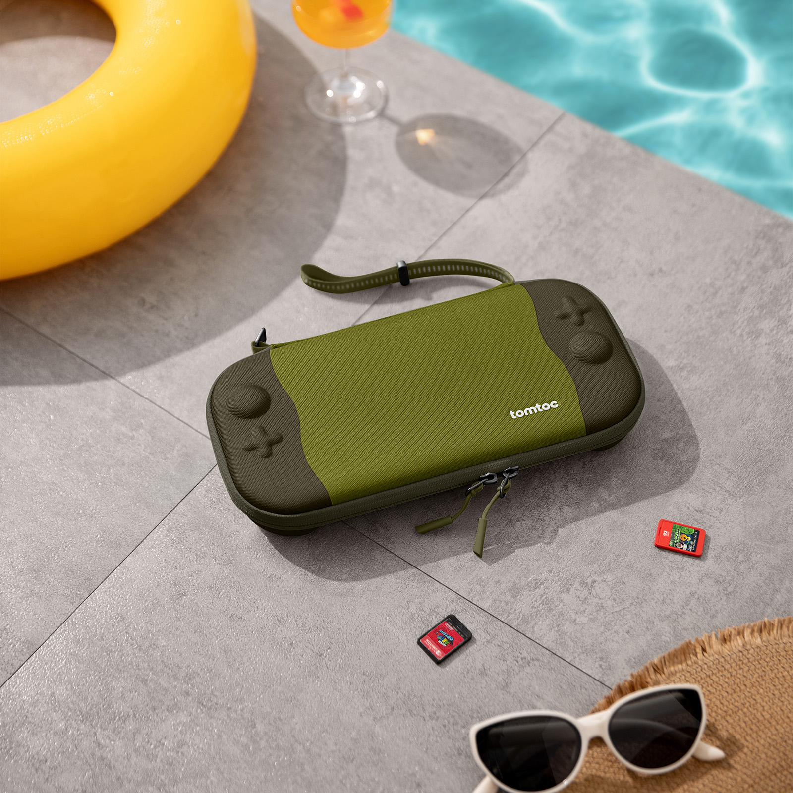 Zielone etui Nintendo Switch leży na patio obok basenu, z żółtym kołem ratunkowym i koktajlem.