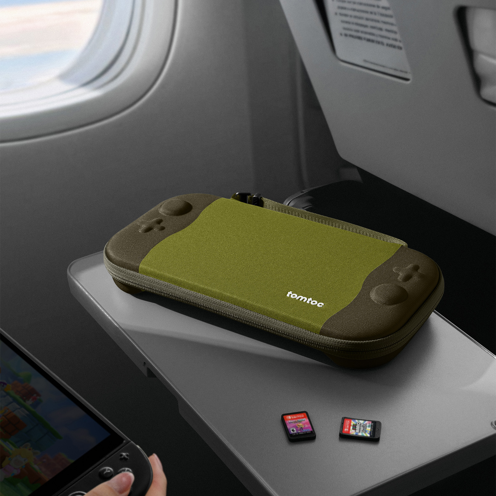 Oliwkowo-zielone etui tomtoc, dwie karty do gier i konsola do gier na stoliku w samolocie obok okna.