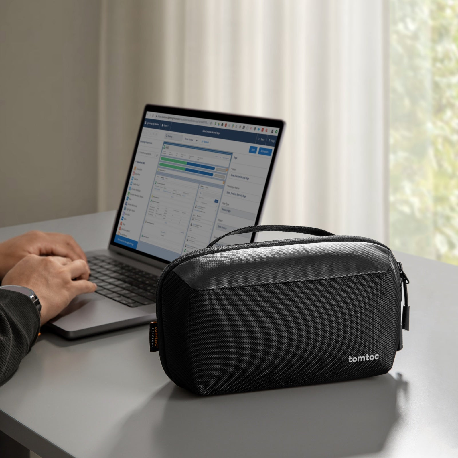 Osoba pisze na laptopie. Czarny futerał z napisem 'tomtoc' na pierwszym planie. Okno w tle.