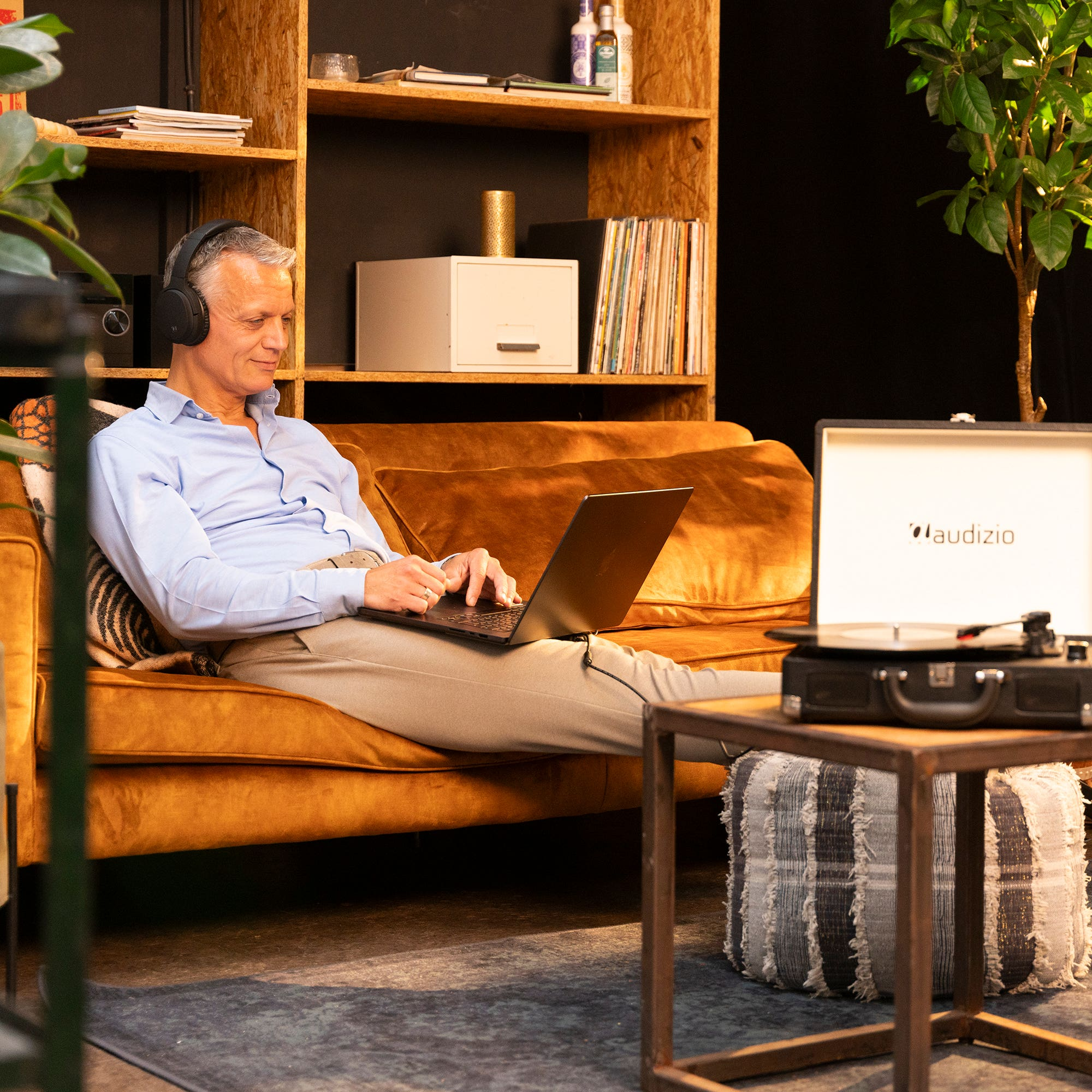 Hombre con auriculares en un sofá usando una laptop. Un tocadiscos está en una mesa. Estanterías y plantas en el fondo.