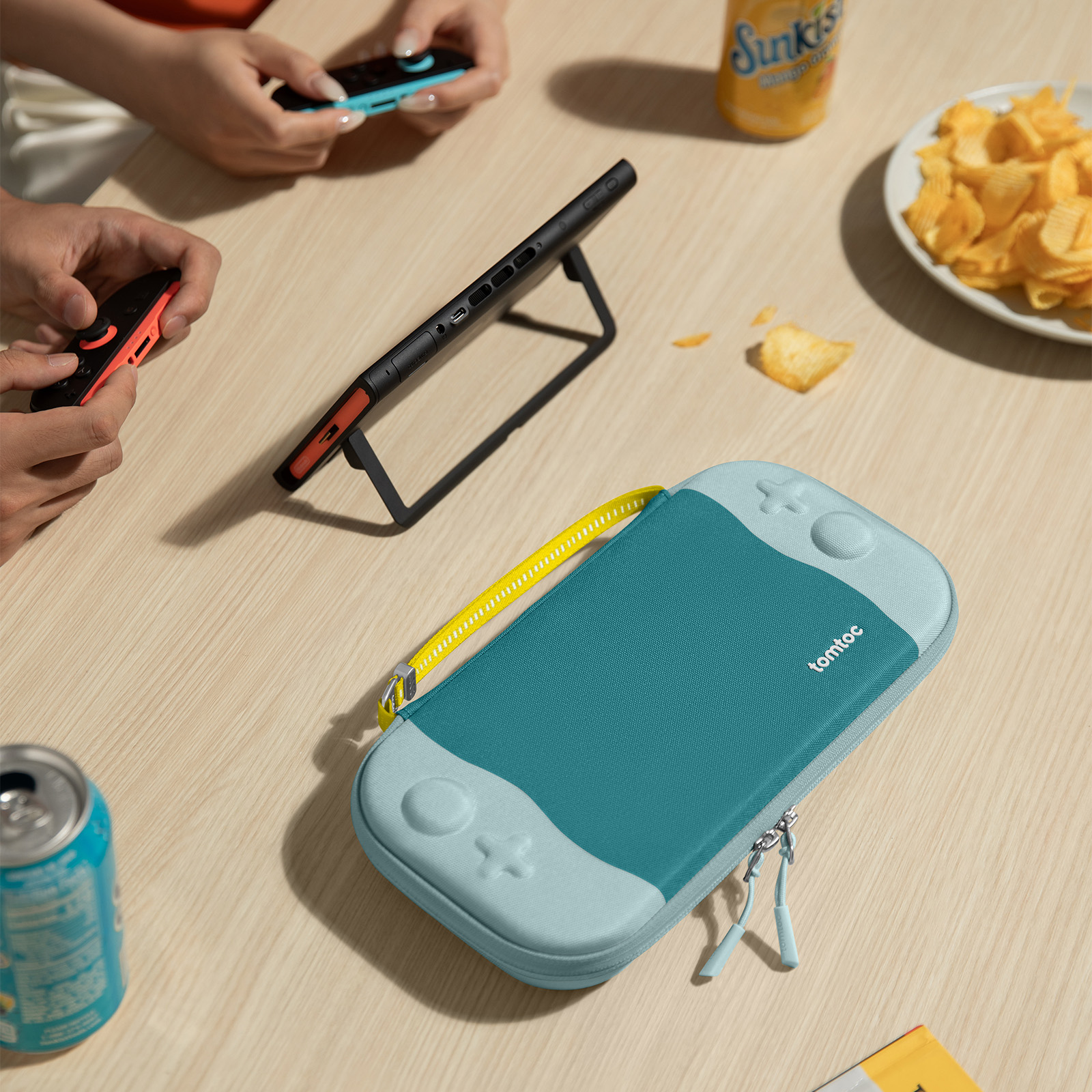 Turkusowe i jasnoniebieskie etui na Switch, dwa kontrolery, puszka sody, chipsy i puszka Sunkist na drewnianym stole.