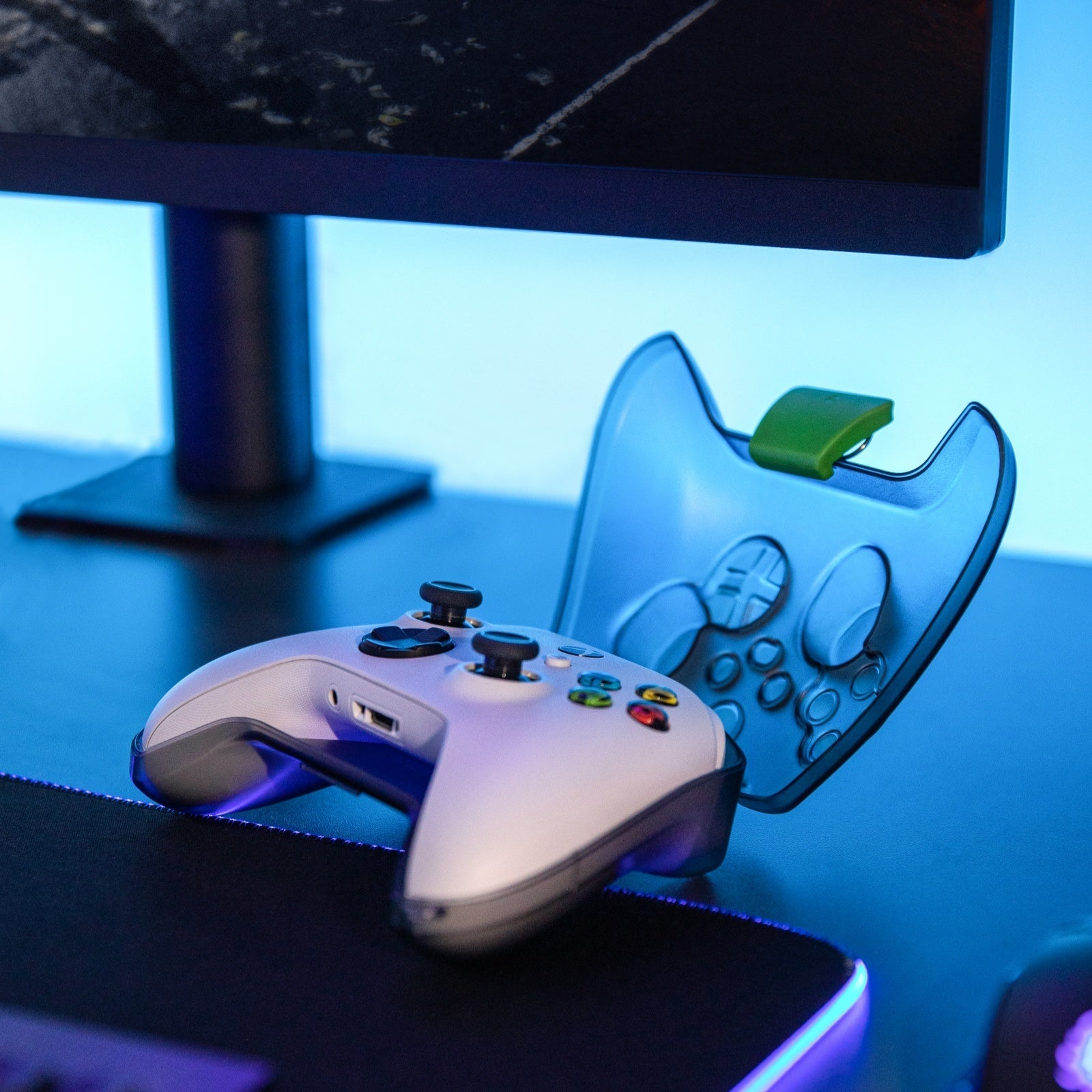 Biały gamepad leży na biurku. Widoczny jest niebieski futerał z zielonym klipsem i stojak na monitor.