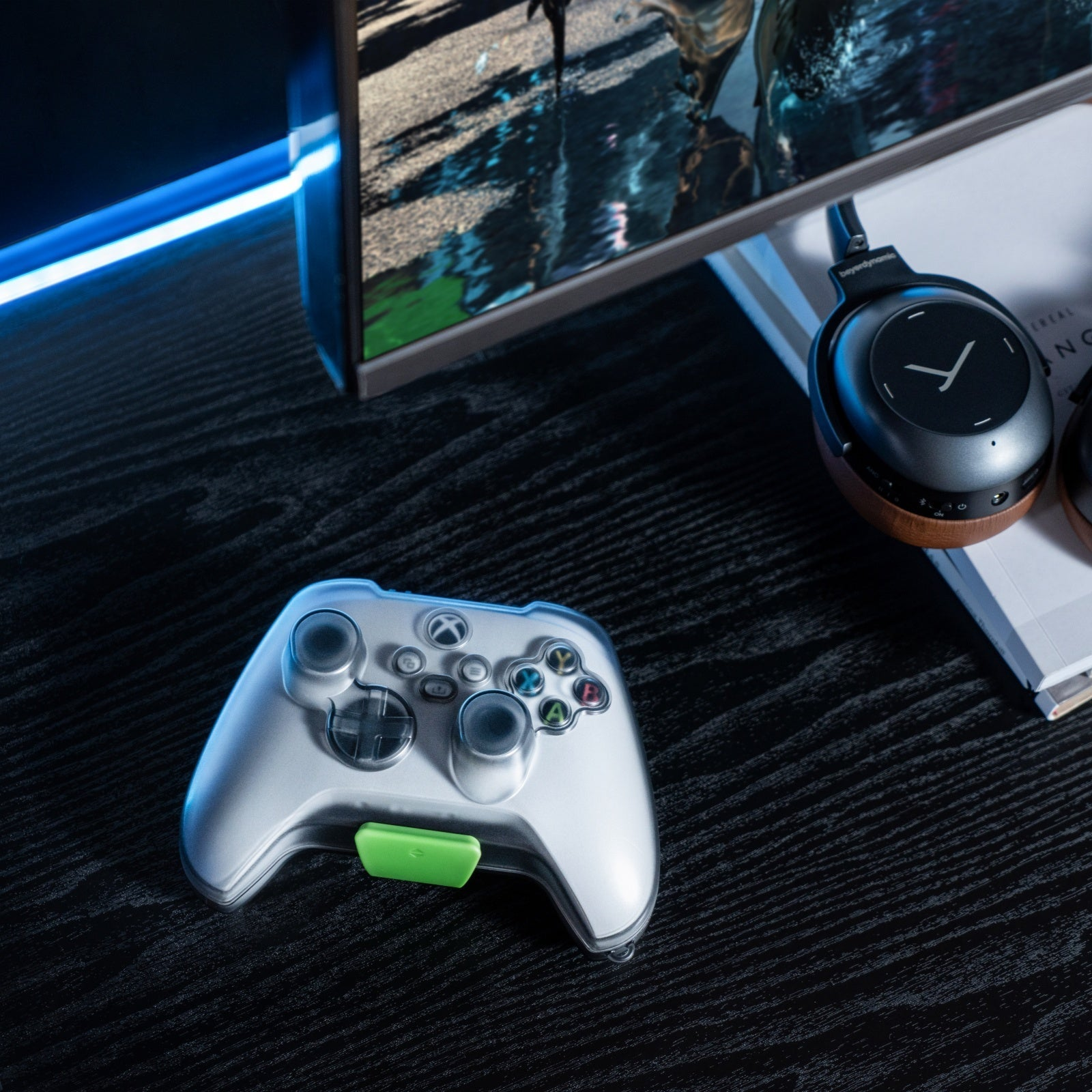 Srebrny kontroler Xbox z zielonym akcentem na czarnym stole. Słuchawki i monitor w tle.