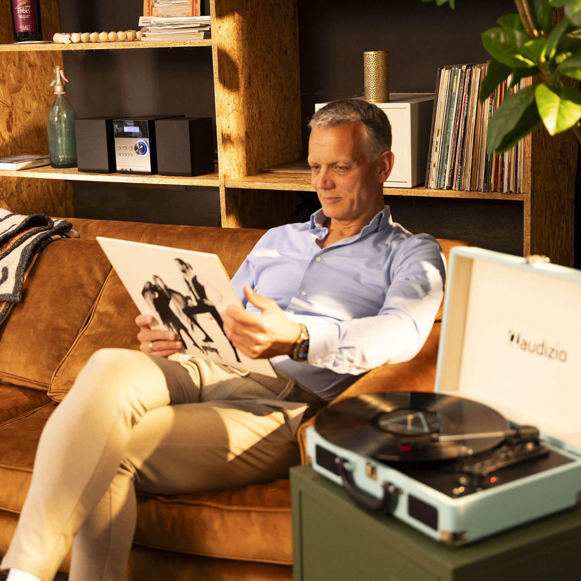 Un hombre sentado en un sofá marrón lee la portada de un disco. Un tocadiscos está a su lado. Detrás de él, estanterías con discos.