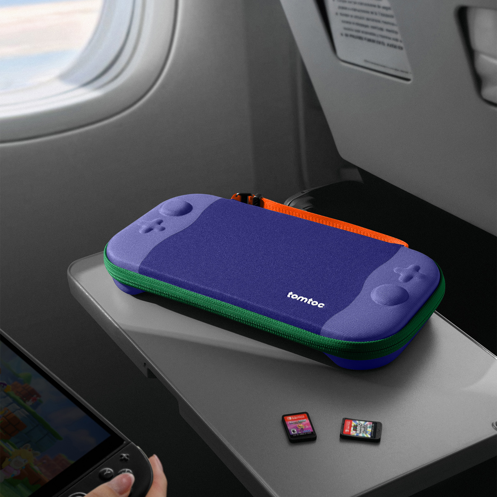 Etui na Nintendo Switch na tacy samolotu. Etui jest fioletowe, zielone i pomarańczowe. Widoczne są dwa kartridże do gier.