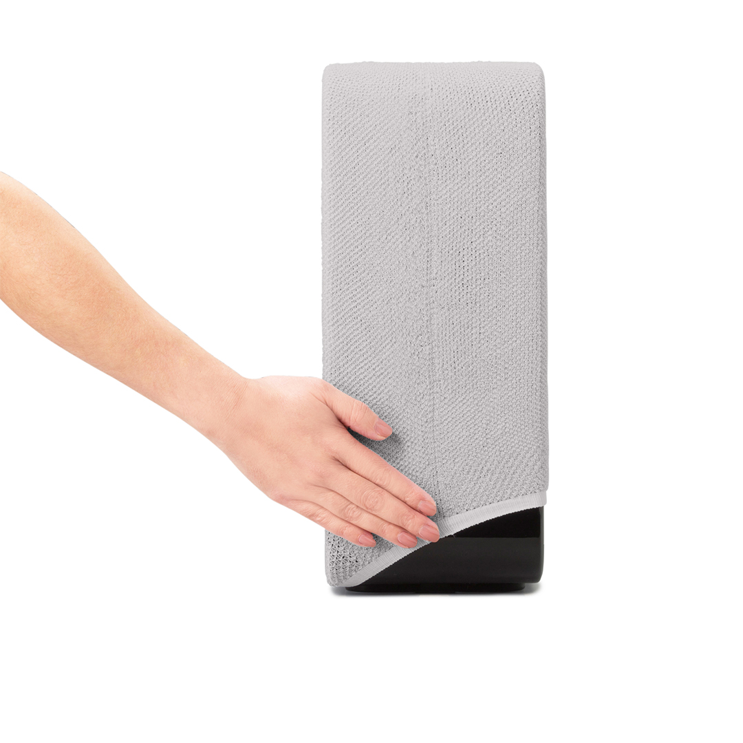 Une main touche un distributeur de serviettes gris. Le distributeur est haut et rectangulaire, sur une base noire. Le fond est blanc.