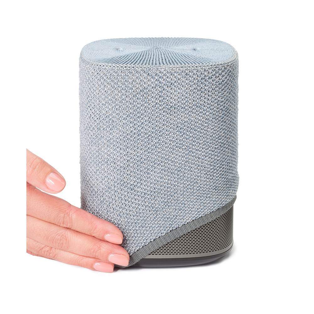 Une main touche un haut-parleur gris. Le haut-parleur a une housse tricotée, un fond en mesh et un dessus plat. Fond blanc.