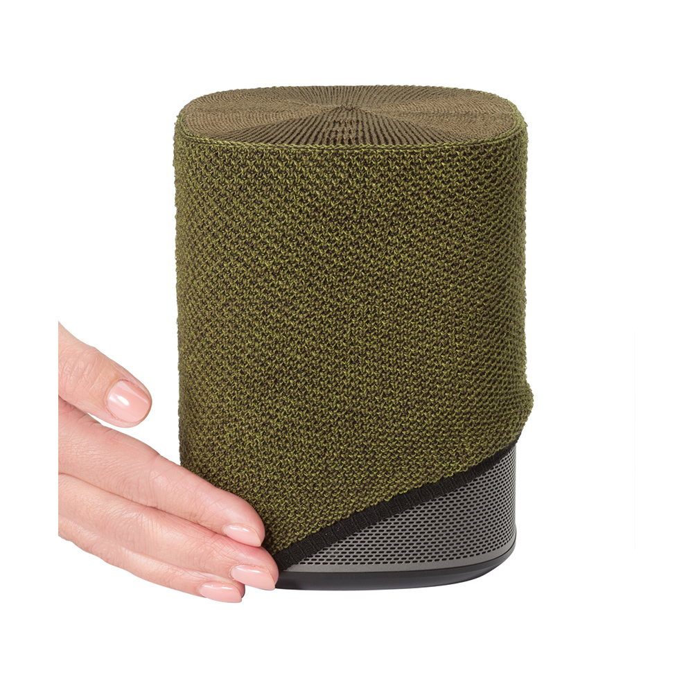 Une main touche un haut-parleur cylindrique. Il a une housse verte tricotée et une base en maille métallique. Le dessus est marron.