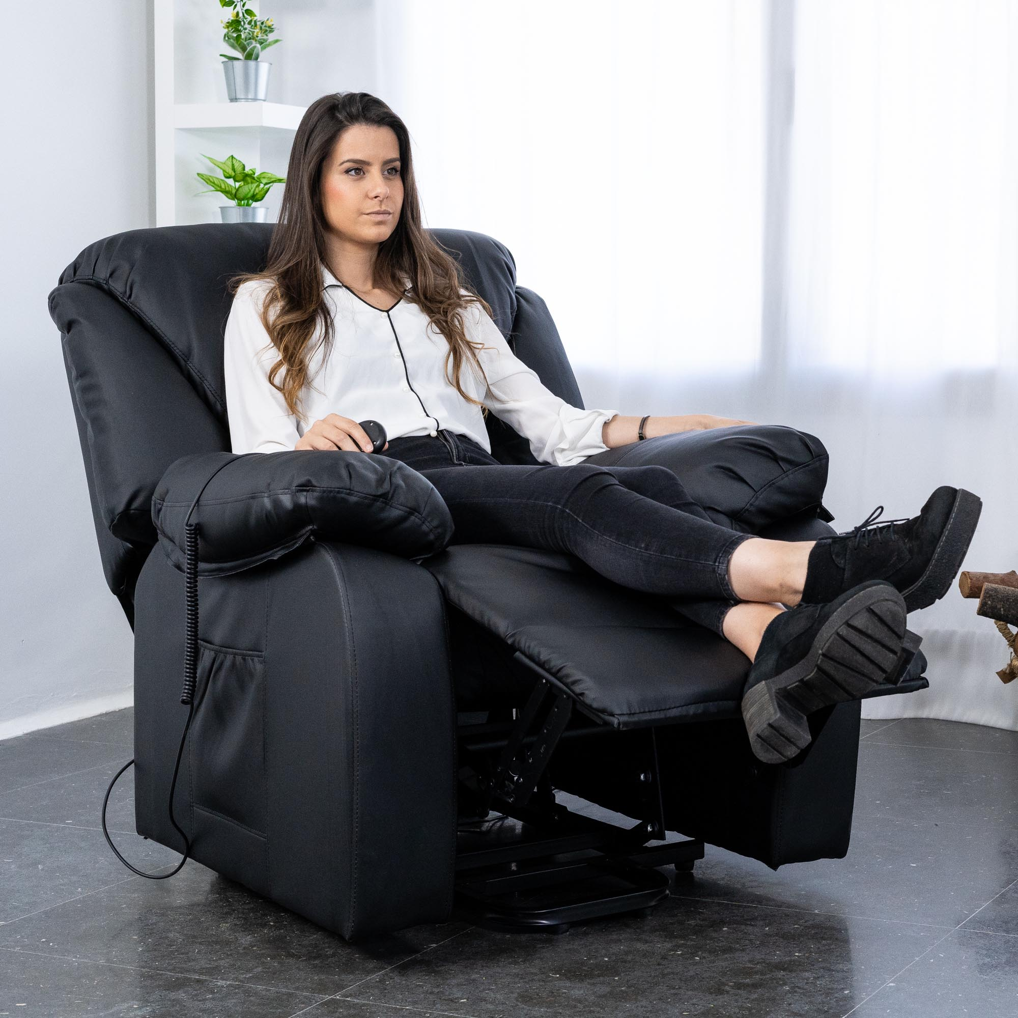 Mujer con ropa negra se relaja en un sillón reclinable negro. Plantas en macetas plateadas están en el fondo, cerca de una ventana.