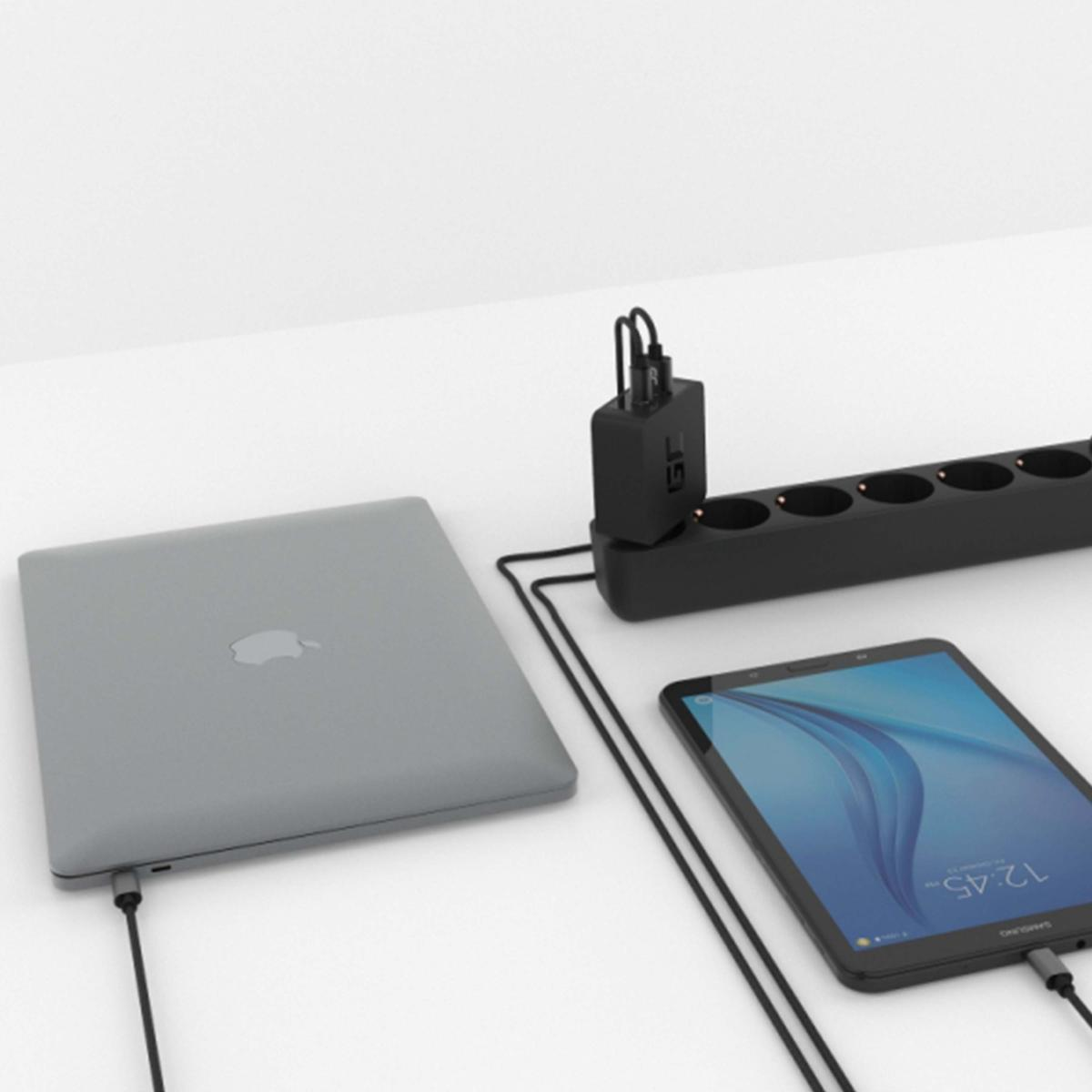 Ein graues Laptop, ein Tablet und eine schwarze Steckdosenleiste mit Ladegerät liegen auf einer weißen Oberfläche. Kabel verbinden die Geräte.