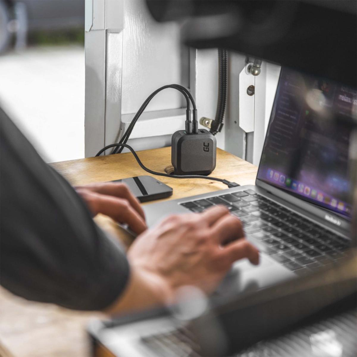 Una persona scrive su un laptop. Un caricatore nero con due cavi è collegato. Telefono e laptop su una scrivania di legno.