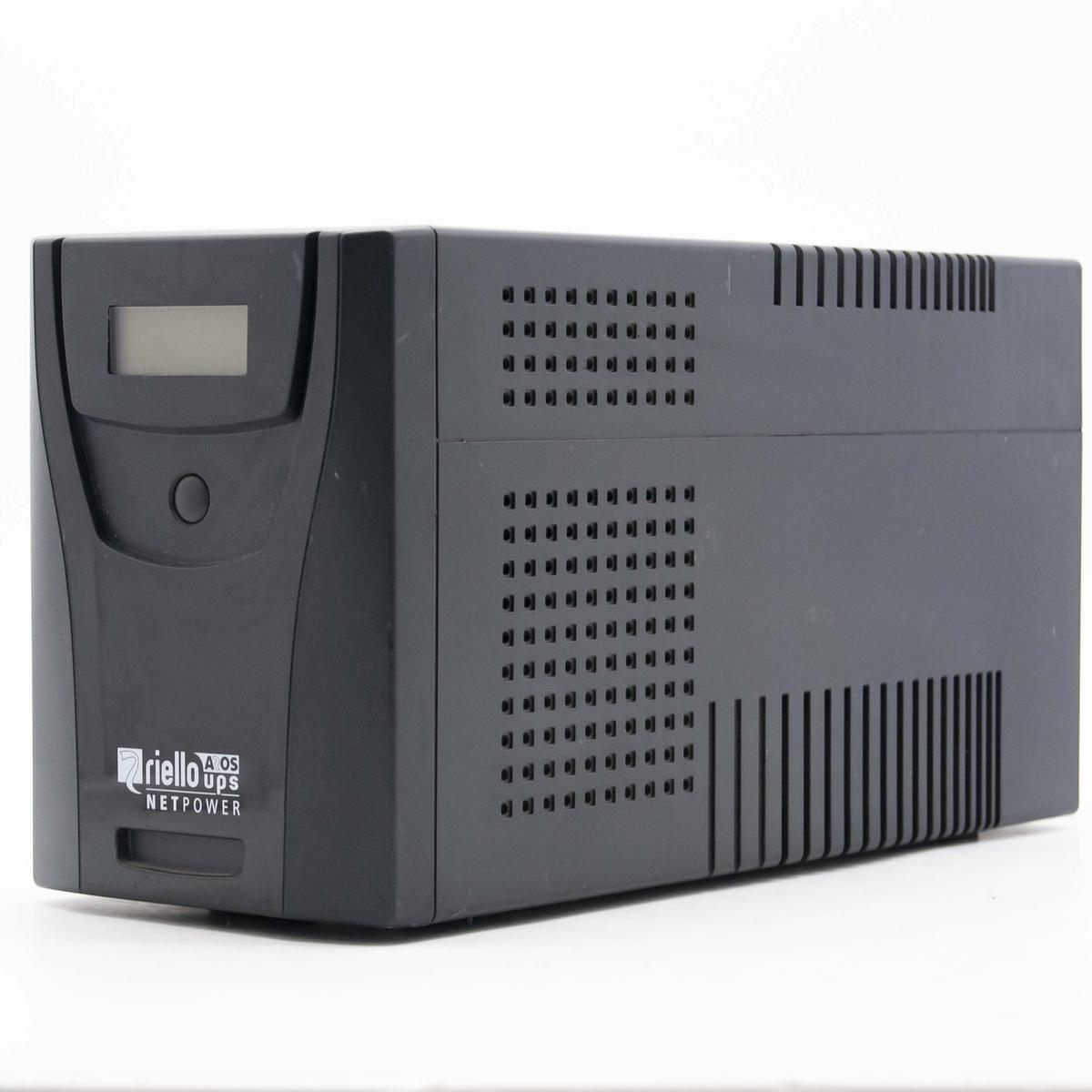 Un SAI Riello Netpower negro. Tiene una pantalla y un botón en la parte delantera. El lateral tiene muchos agujeros cuadrados para la ventilación.