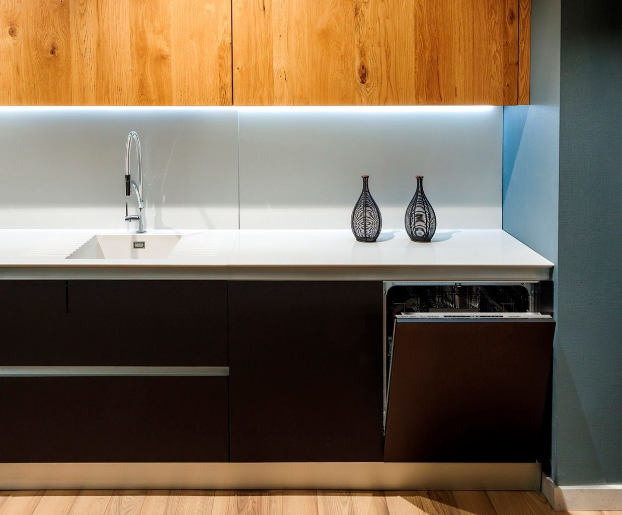 Cocina moderna con armarios de madera, encimera blanca, fregadero, lavavajillas y jarrones decorativos. Se ven armarios marrones y una pared azul.