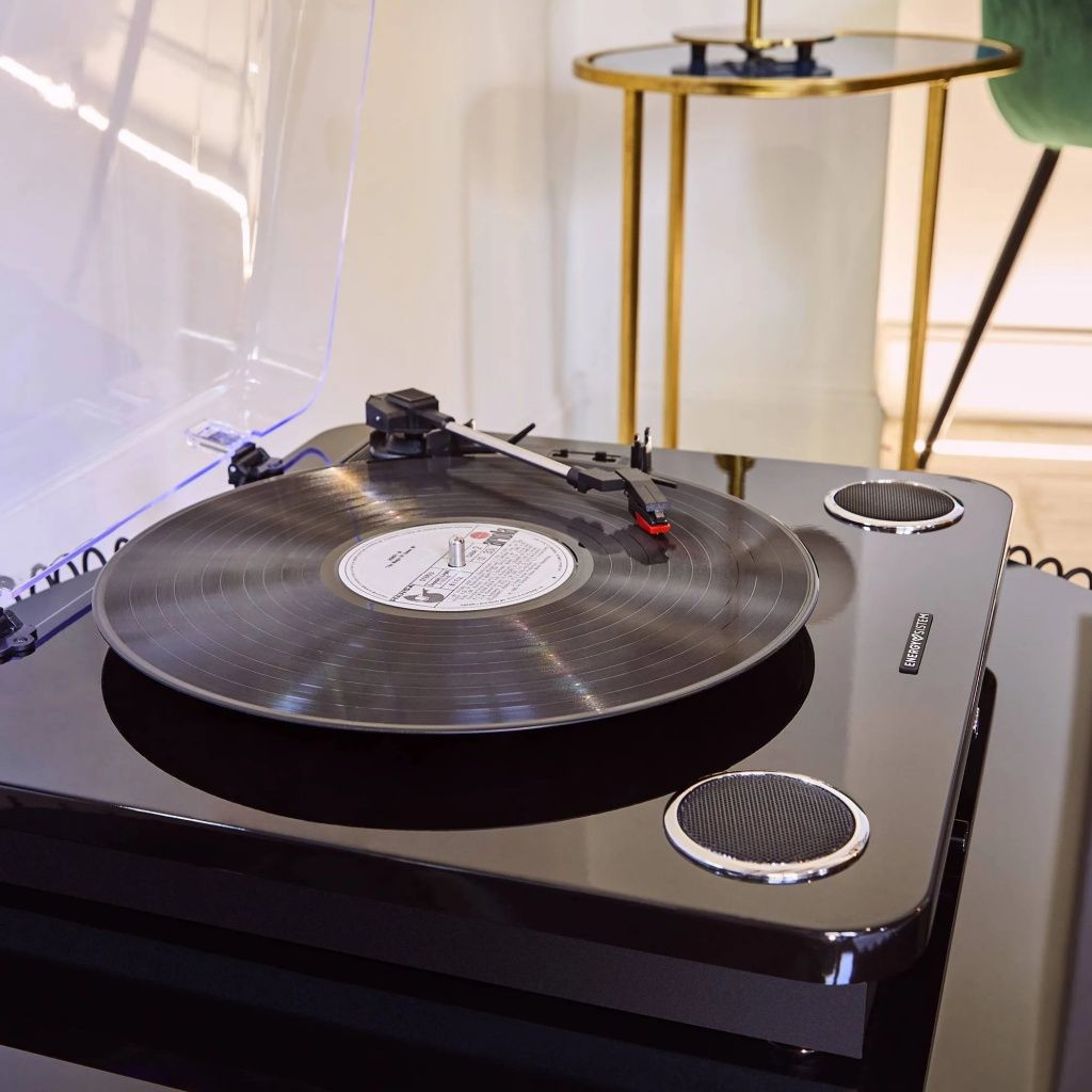 Un tocadiscos negro con un disco de vinilo está reproduciendo música. Una mesa pequeña y una silla verde están en el fondo.