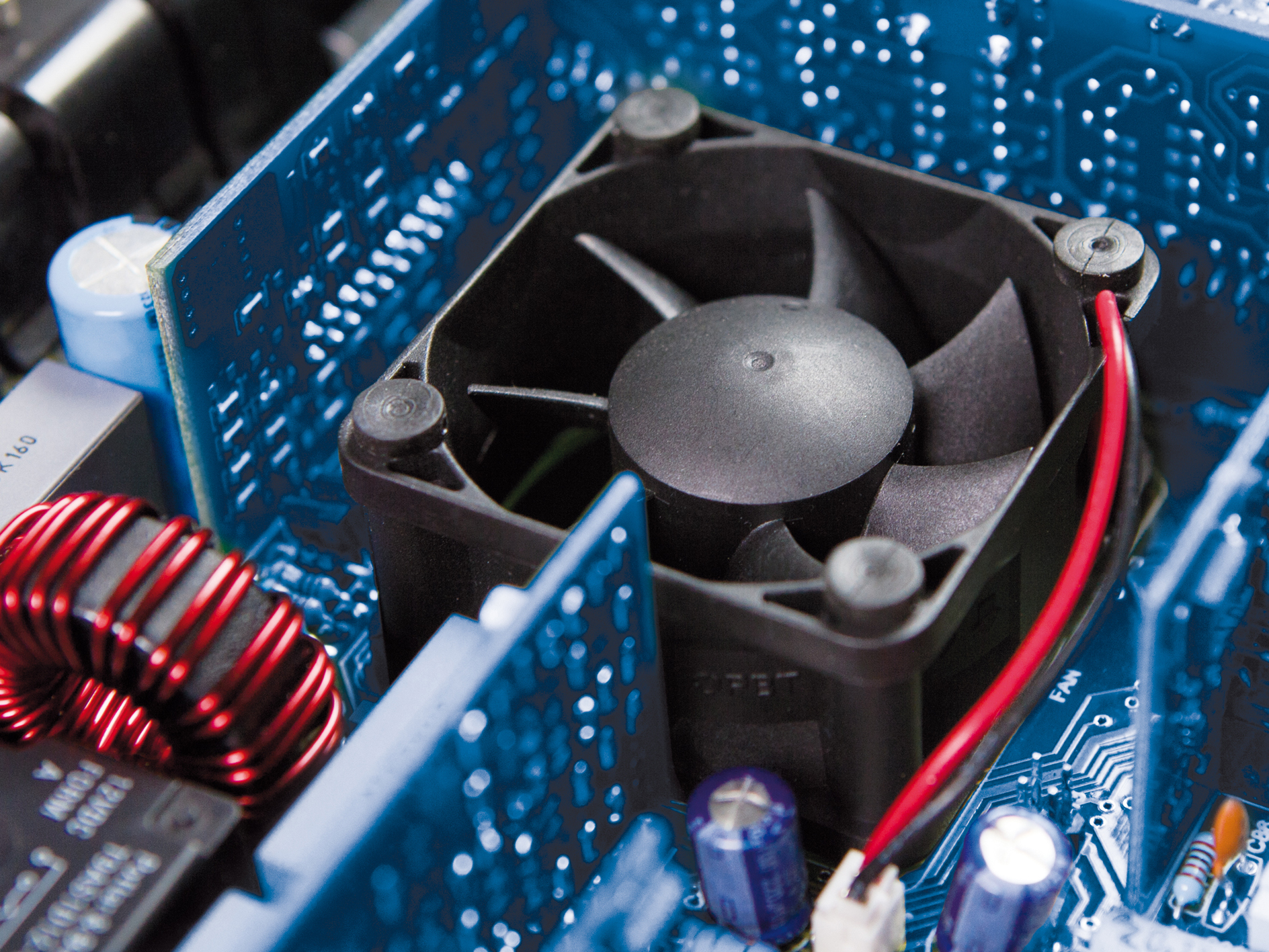 Nahaufnahme einer Computerplatine. In der Mitte befindet sich ein schwarzer Lüfter. Rote und schwarze Drähte sind sichtbar. Blaue Komponenten umgeben ihn.