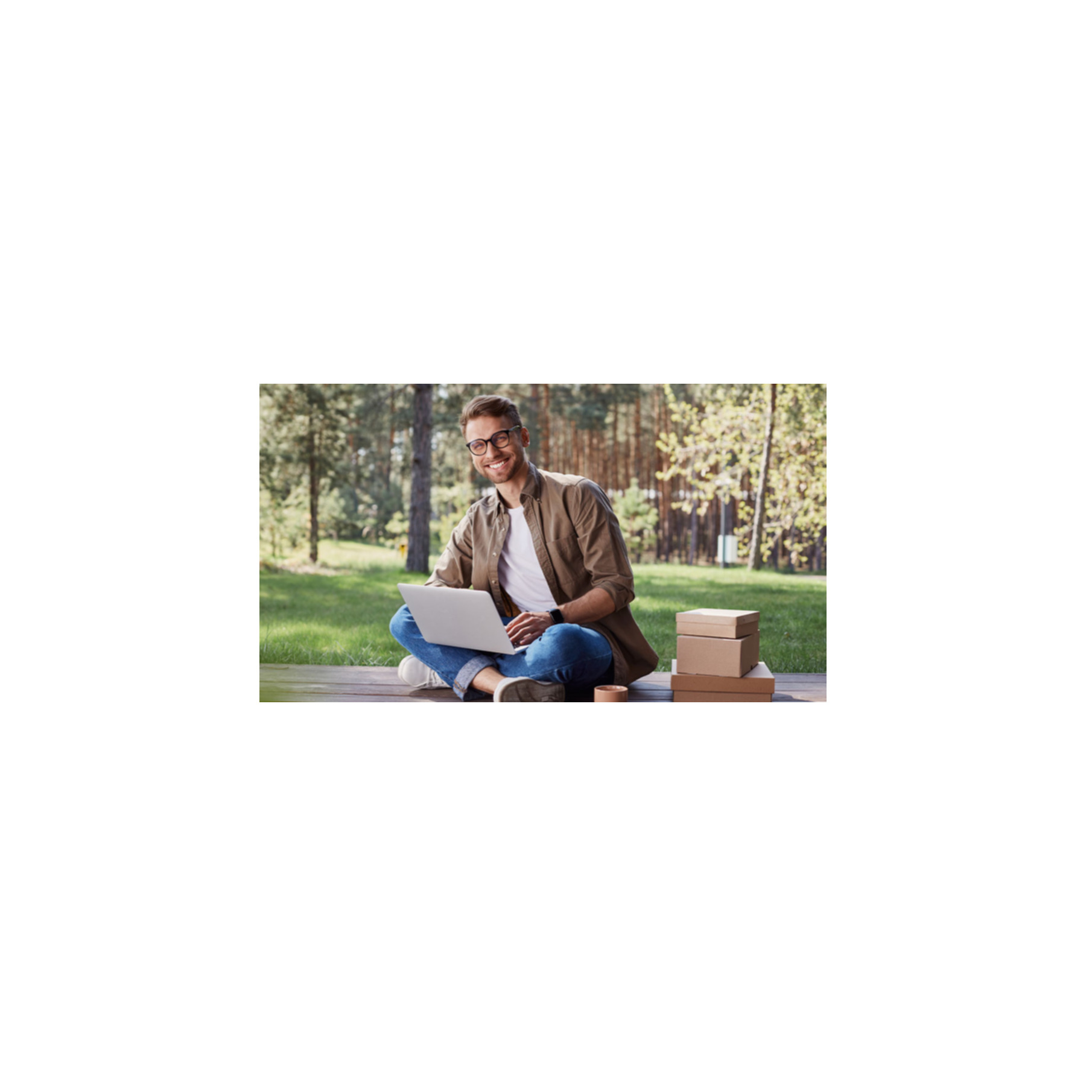 Mężczyzna w okularach siedzi ze skrzyżowanymi nogami, używając laptopa na zewnątrz. Pudełka są ułożone w pobliżu. Drzewa i trawa w tle.