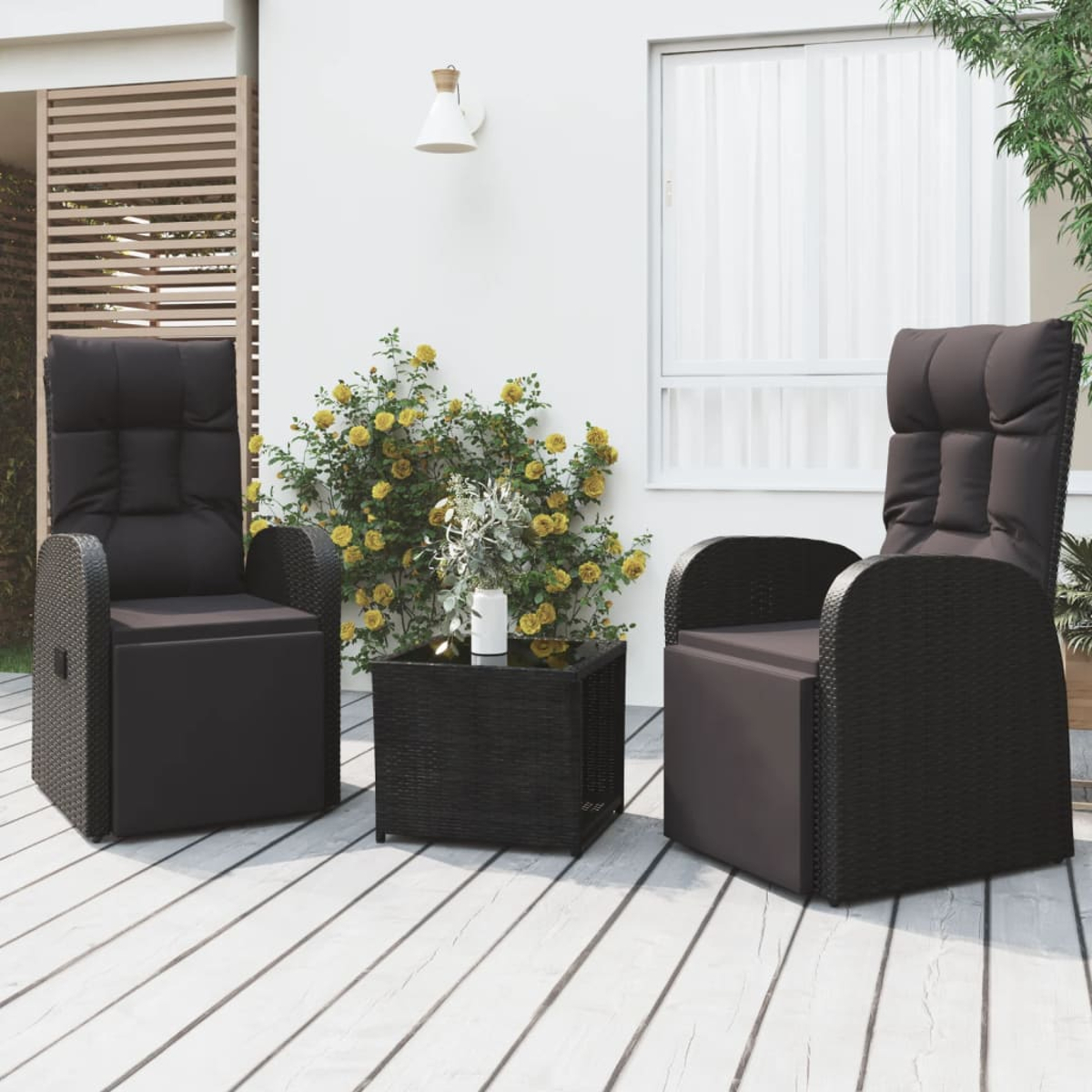 Zwei schwarze Korbstühle mit Kissen und ein kleiner Tisch auf einem Holzdeck. Gelbe Blumen und weiße Wand im Hintergrund.