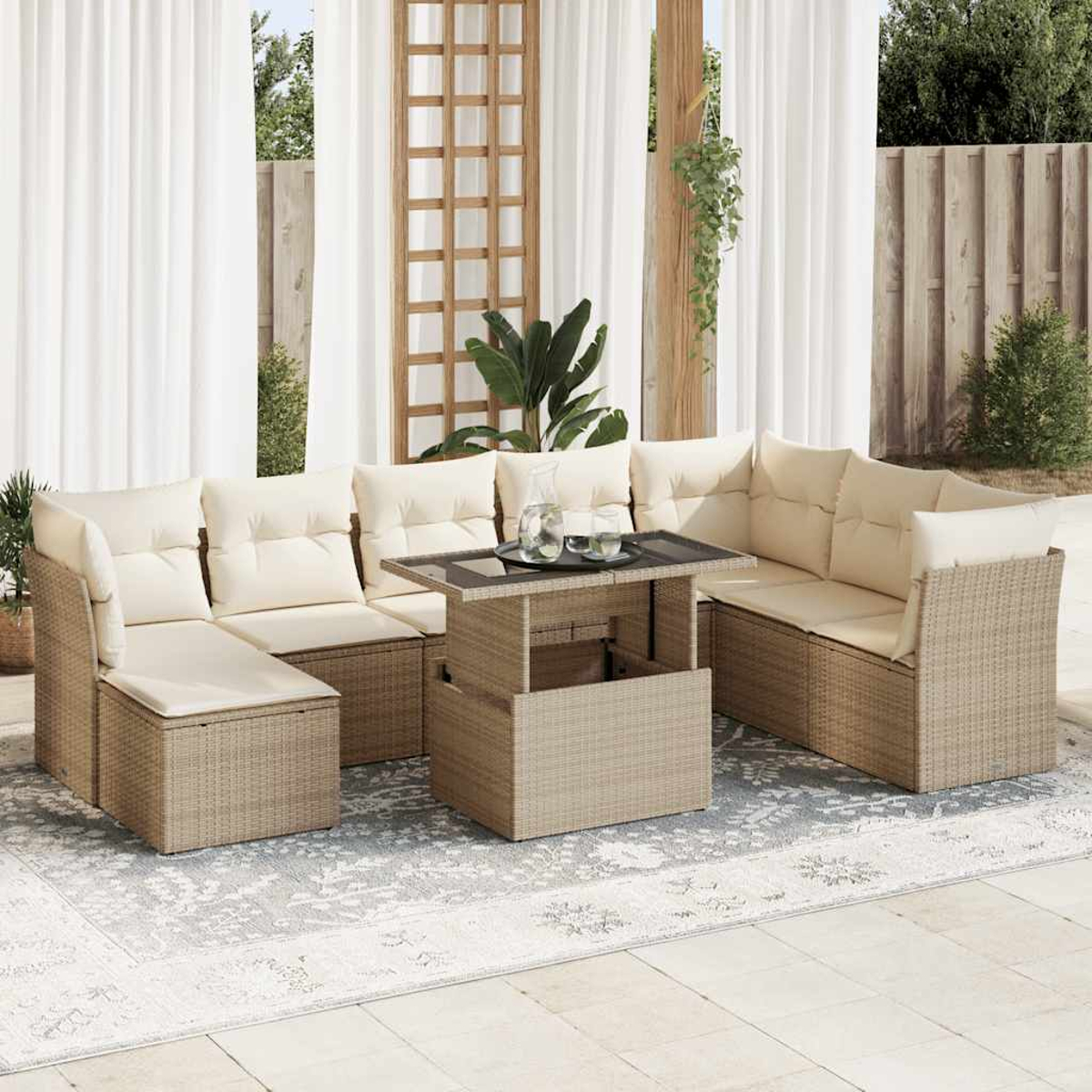 Divano componibile in rattan da esterno con cuscini color crema e tavolo. Tappeto beige su un patio. Tende bianche e staccionata in legno sullo sfondo.