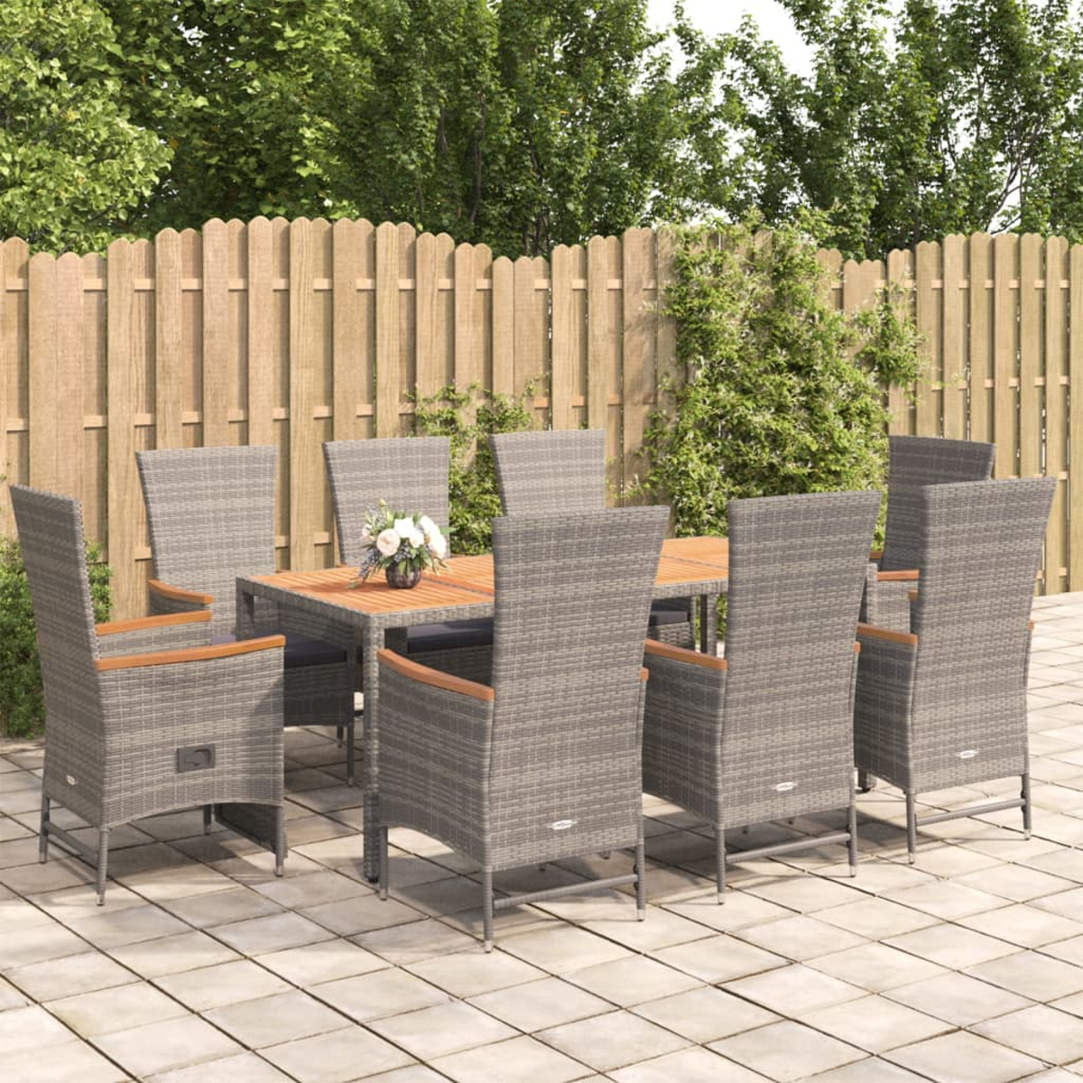 Essgruppe im Freien mit sechs grauen Korbstühlen und einem Holztisch. Auf einer Terrasse mit Holzzaun und grünen Bäumen im Hintergrund.