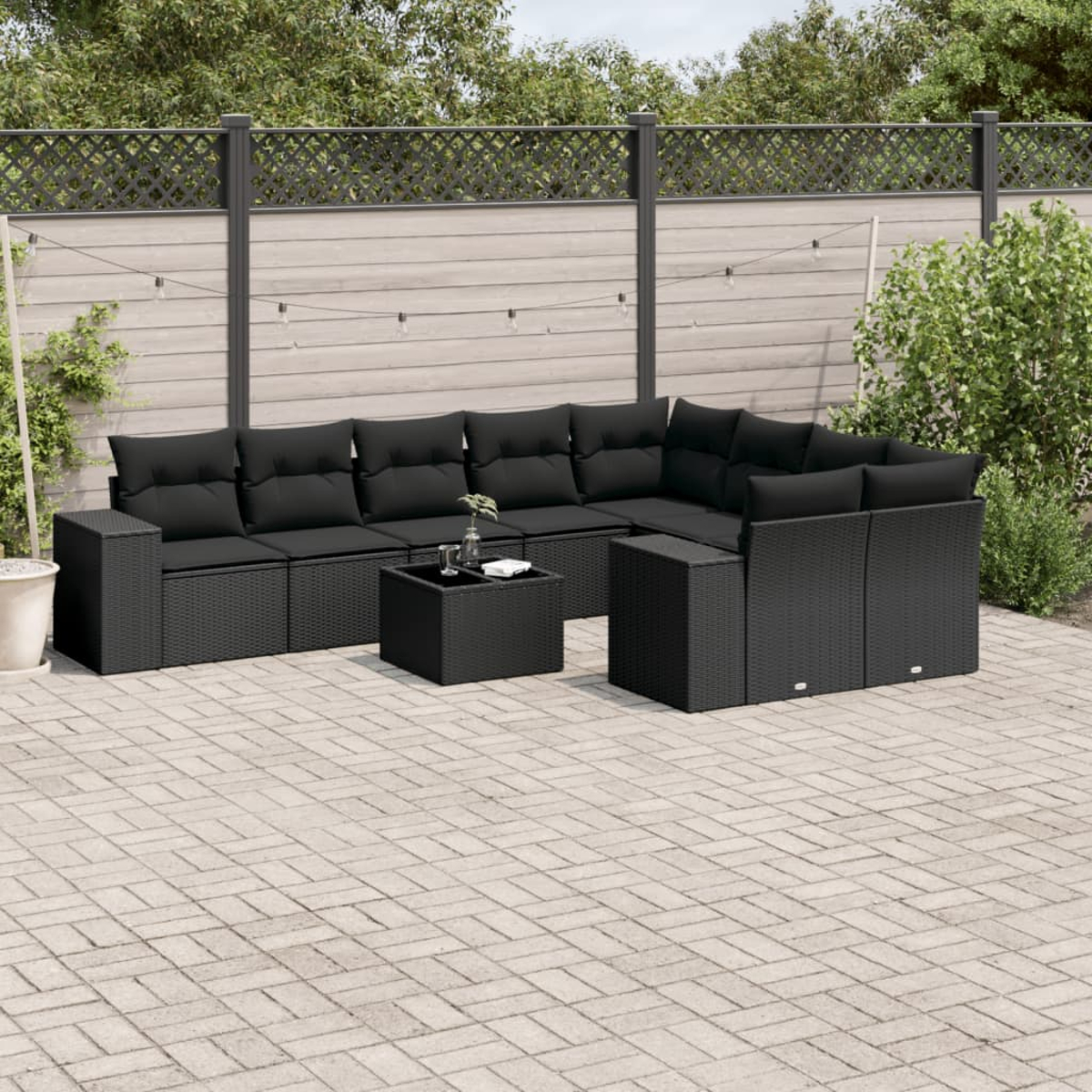 Schwarzes Ecksofa mit Kissen und quadratischem Couchtisch auf einer Ziegelterrasse. Im Hintergrund ein Zaun und Grün.