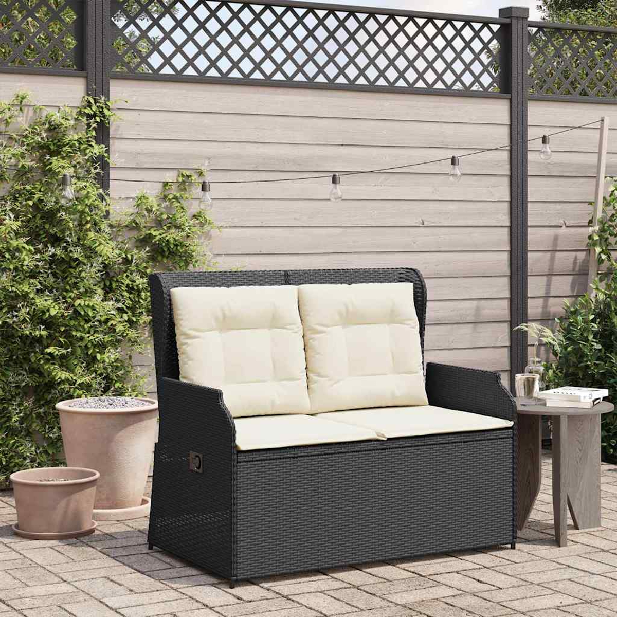 Una panca nera in vimini con cuscini color crema si trova su un patio. Un tavolino è vicino con un bicchiere e libri. Una recinzione di legno è sullo sfondo.