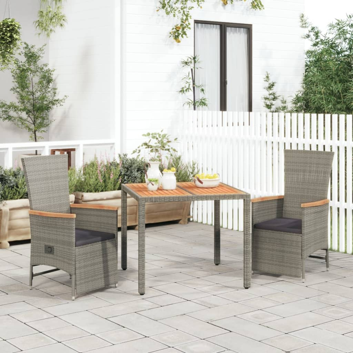 Szene auf der Terrasse: graue Stühle, ein Tisch mit Obst und Pflanzen. Weißer Zaun und Gebäude im Hintergrund. Gepflasterter Boden.