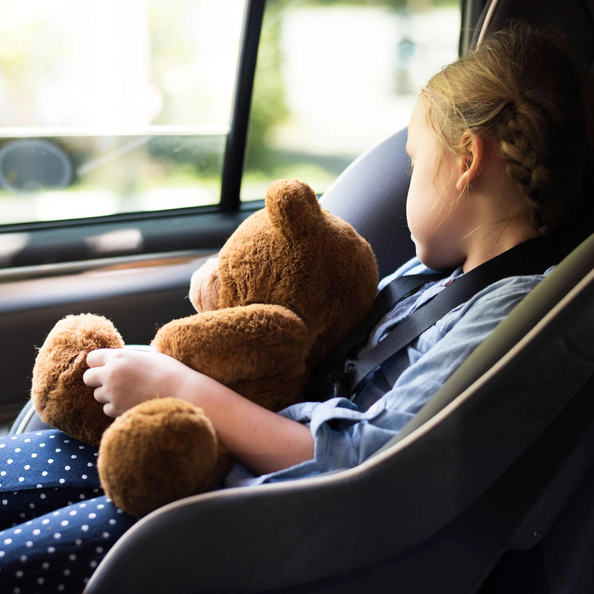 Ein Kind im Autositz hält einen Teddybären. Das Kind trägt ein blaues Hemd und gepunktete Hose. Draußen sind Bäume zu sehen.