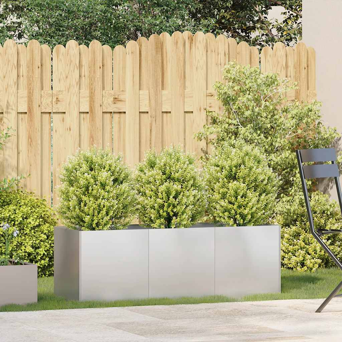 Scena di giardino con recinzione in legno, tre cespugli verdi in una fioriera argentata e una sedia nera a destra. Arbusti verdi.
