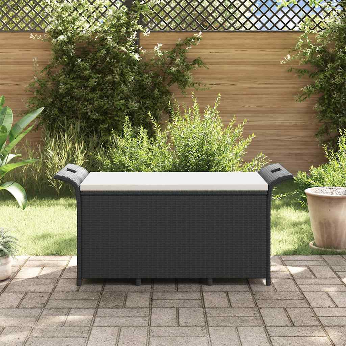 Eine schwarze Aufbewahrungsbank mit weißem Kissen steht auf einer Terrasse. Grüne Pflanzen und ein Holzzaun befinden sich im Hintergrund.