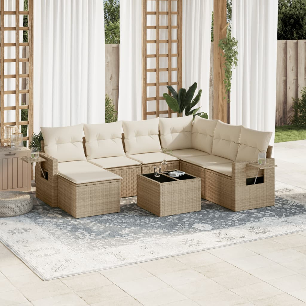 Area salotto all'aperto con cuscini beige e mobili in vimini. Include un tappeto, un tavolino e tende. Piante sullo sfondo.