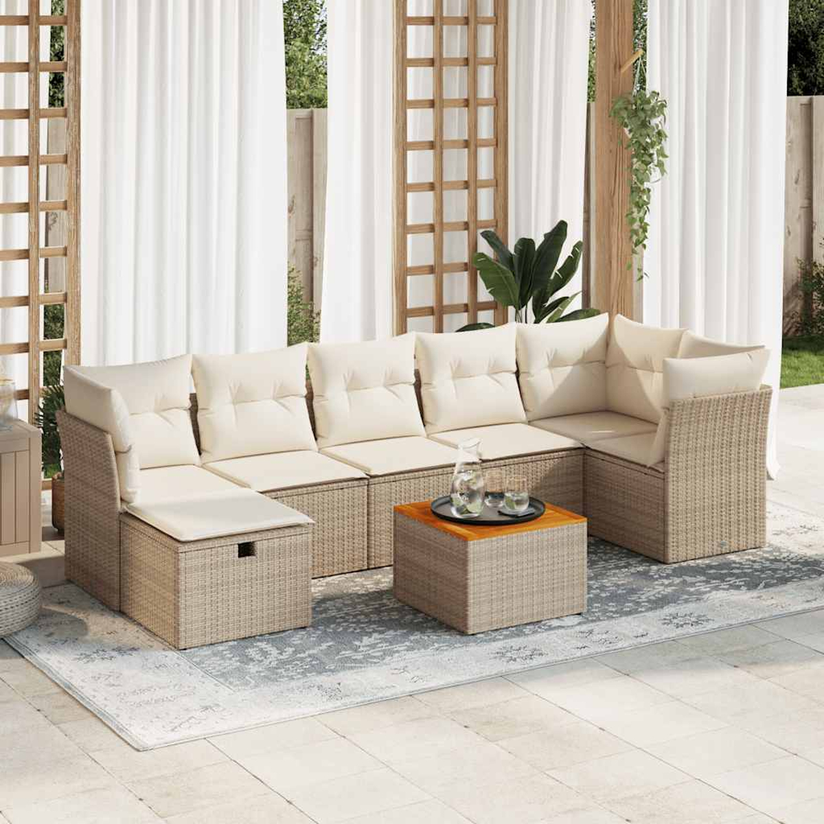 Divano angolare da esterno con cuscini beige e base in rattan. Un tavolino quadrato con bevande davanti. Tende bianche e piante sullo sfondo.